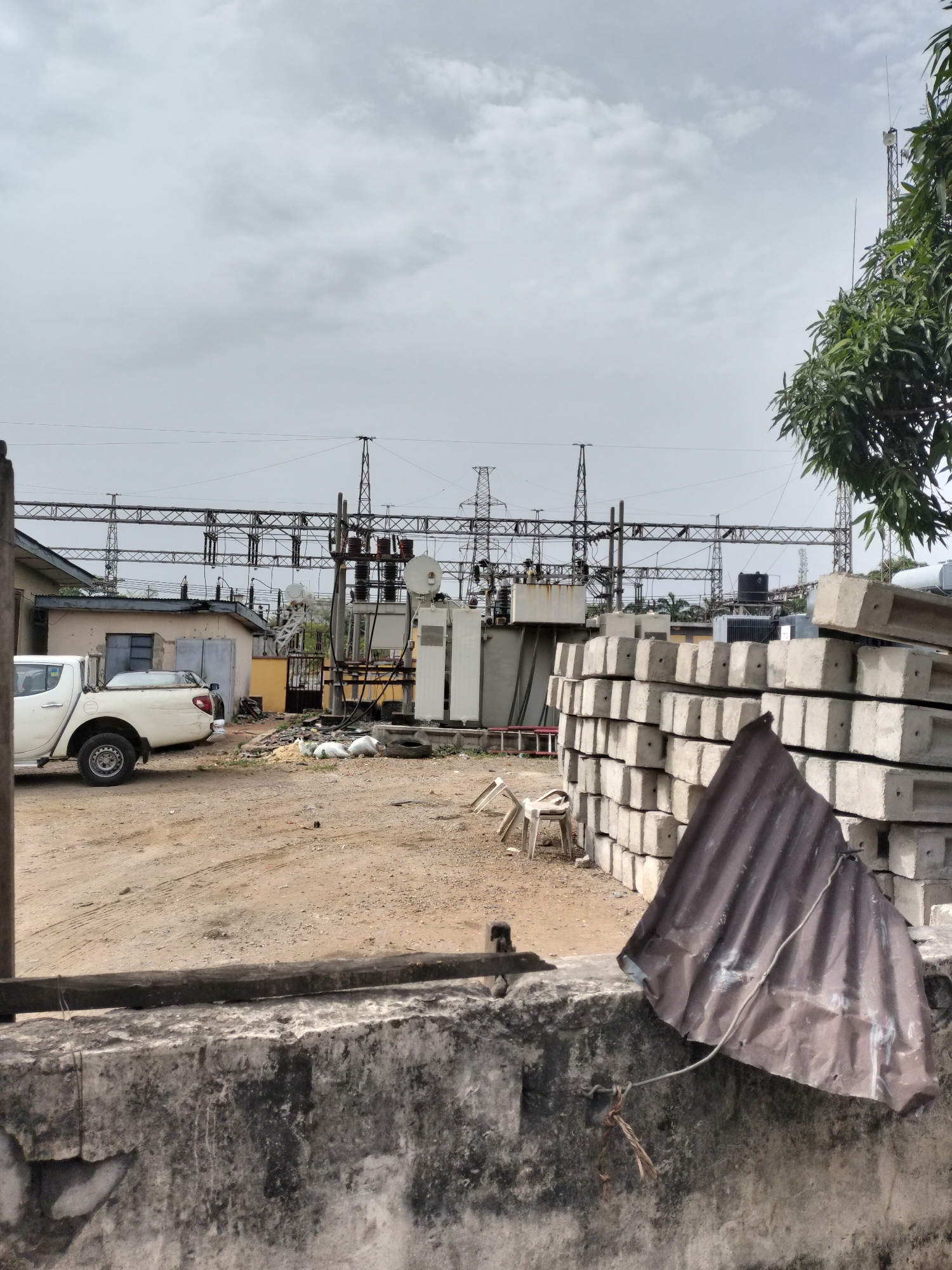 The image depicts an outdoor scene, likely an industrial or utility area in Lagos, Nigeria. In the background is a high-voltage electrical substation with transformers, switchgear, and supporting structures. Power lines extend across the top of the frame. To the left, a white pickup truck is parked near a small building. The foreground features a weathered concrete wall with a piece of corrugated metal leaning against it. A stack of precast concrete blocks is visible behind the metal sheet. Several white plastic chairs are scattered on the dirt ground. The sky is overcast.