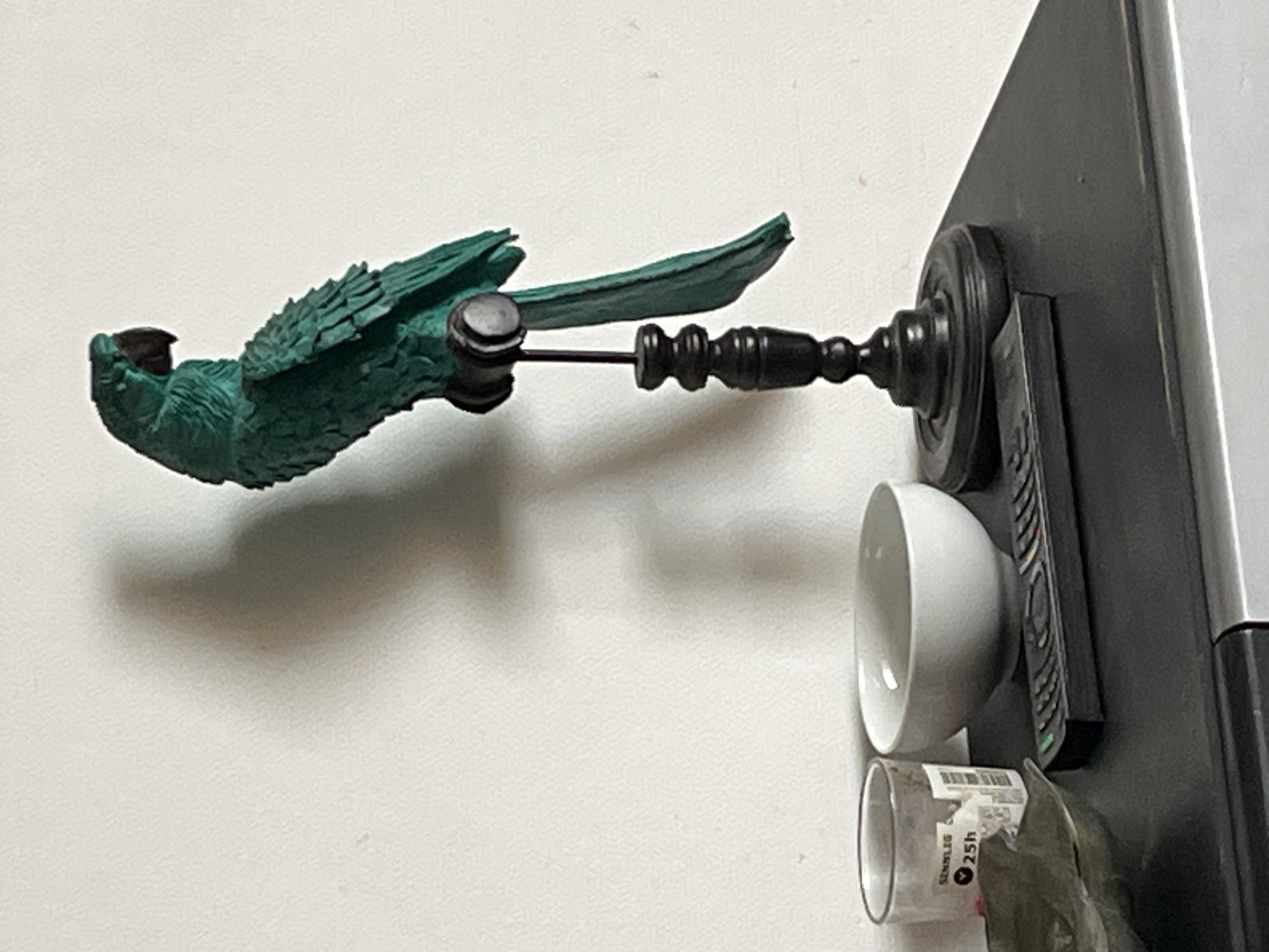 A green parrot statue is attached to a black, ornate decorative object which is affixed to the side of a black refrigerator. A small white bowl and a clear glass jar with a "Sennlig" label are also attached to the refrigerator's side. The setting appears to be an interior space, possibly a kitchen. The location in Palavas-les-Flots, France, is unknown as there are no clues to identify a particular place.