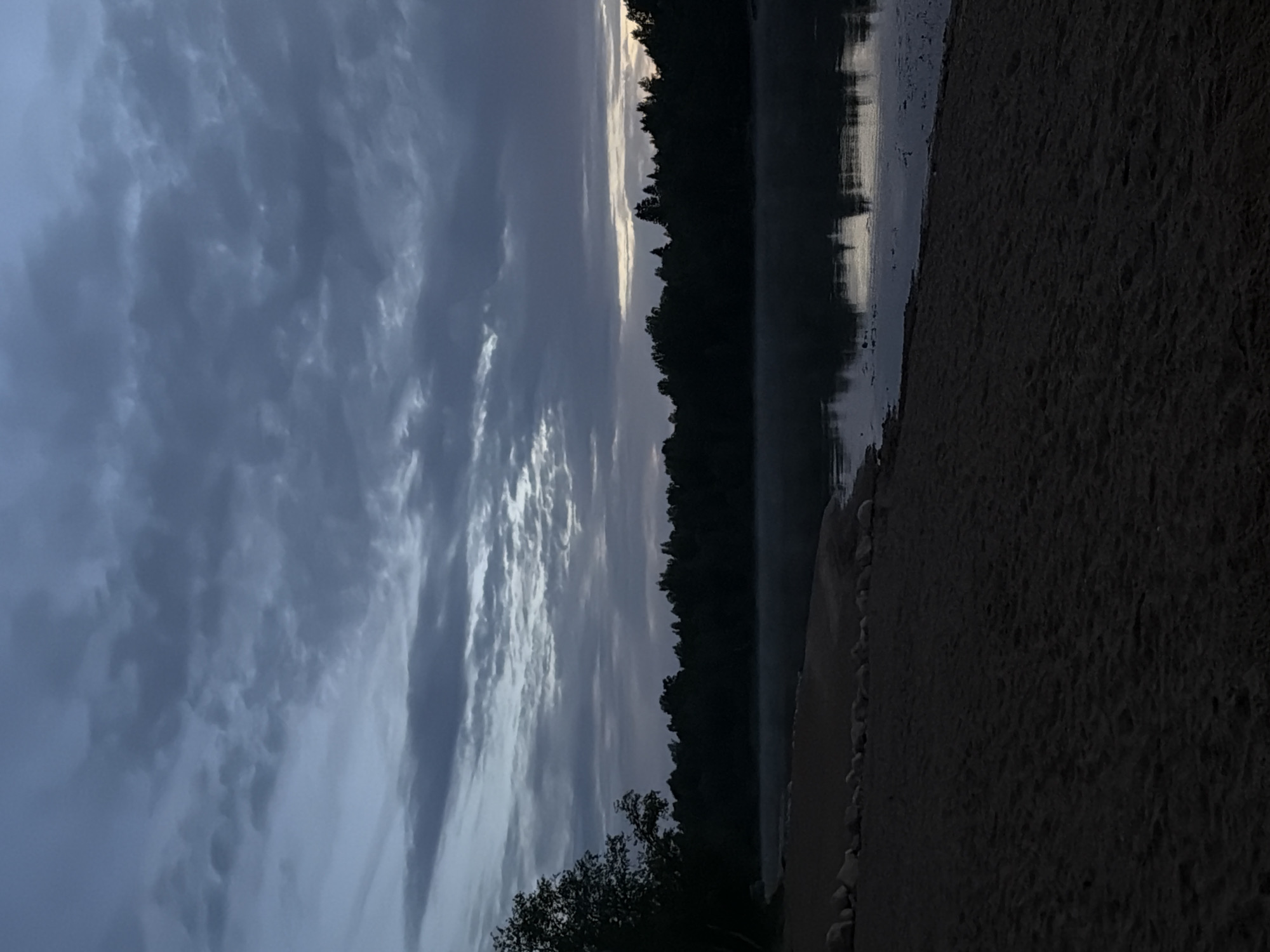 The image depicts a serene twilight scene at the shore of Lake Verkhneye in Solov'evo, Russia. A dark, sandy beach occupies the foreground, with a line of stones marking the transition to the water's edge. The lake itself is calm, reflecting the muted colors of the sky and the dense, dark silhouette of a forest that lines the opposite bank. A faint mist hovers over the water's surface, adding to the tranquil atmosphere. The sky above is filled with dramatic, layered clouds in shades of gray and blue, hinting at the end of the day or the approach of dawn. No people or specific actions are visible in the scene, emphasizing the quiet and natural beauty of the location.