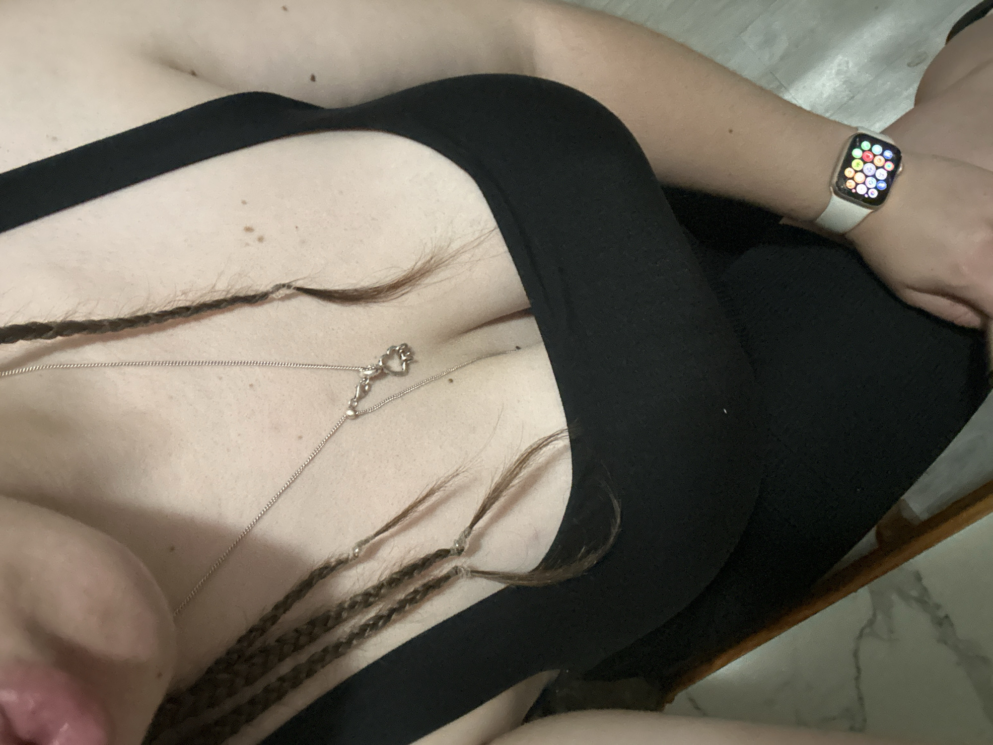 A close-up, overhead shot shows a person wearing a black tank top and a silver necklace. The person has braided hair. On their left wrist, they are wearing a white Apple Watch. The background appears to be an indoor setting with a marble-patterned floor and a wooden chair. There is no indication of the city of Basarabeasca, Moldova.