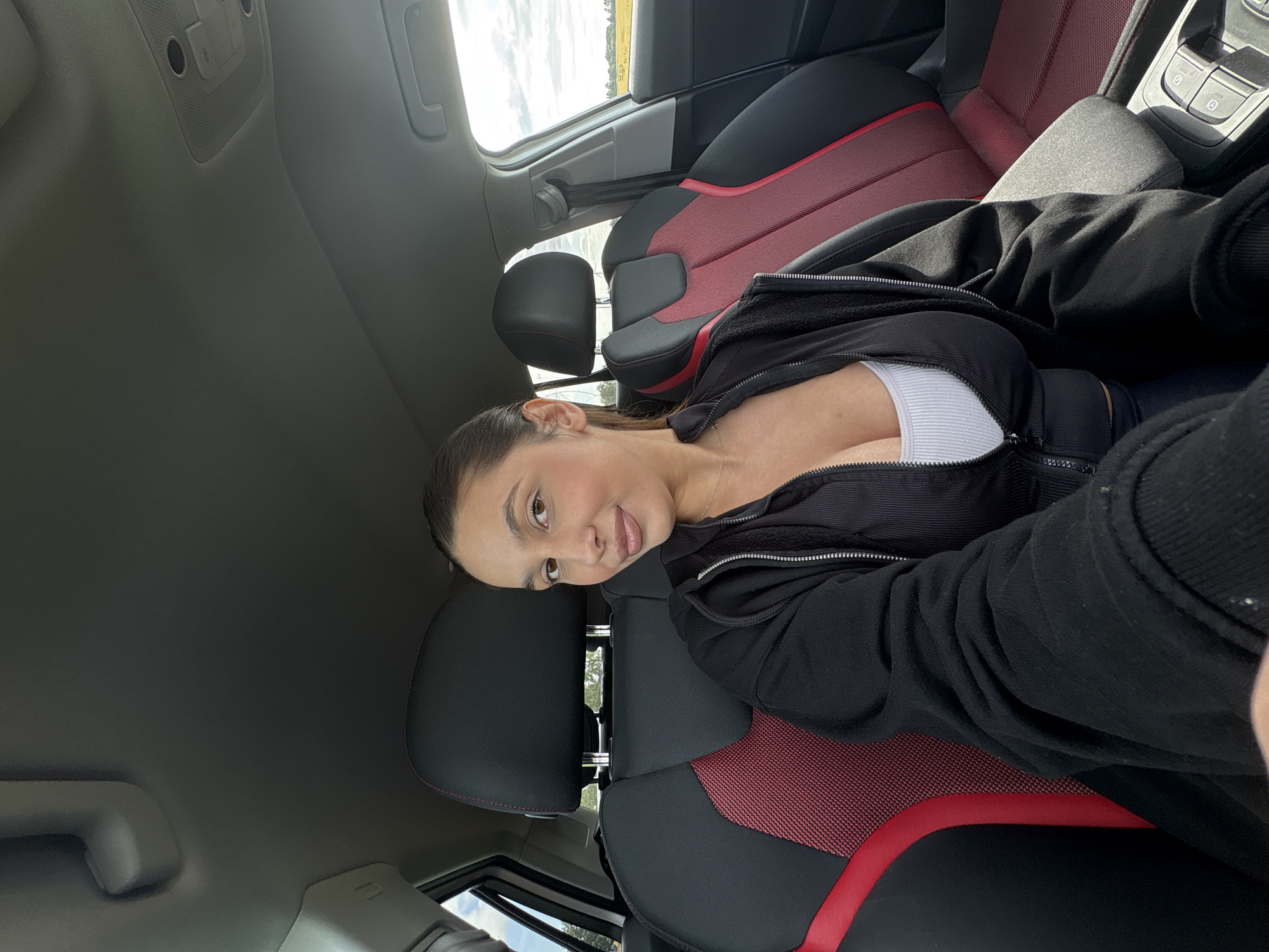 A light-skinned young adult female with dark hair pulled back and light brown eyes is seated in the front passenger seat of a car, taking a selfie. She wears a white ribbed tank top beneath a partially unzipped black athletic-style top, further covered by a black hoodie. A slender silver necklace is visible. The vehicle's interior features black seats with red fabric inserts and red stitching. Through the car windows, an outdoor scene depicting open fields under a cloudy sky is visible. The provided context indicates the location is in Wegberg, Germany, though this specific geographical detail is not verifiable from the image itself.