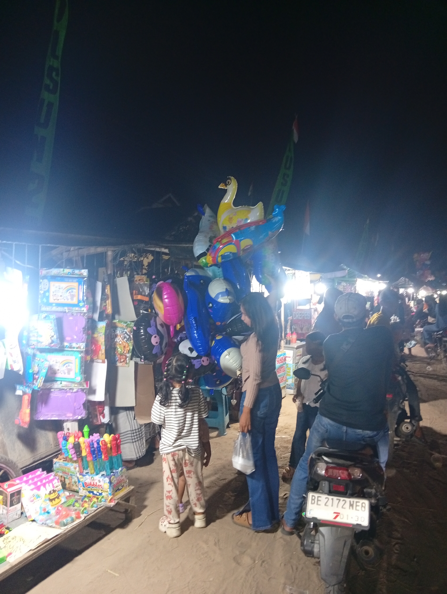 This is a night time, low-angle, wide shot of a street market in Sri Gading, Indonesia. A young girl, wearing a striped shirt and patterned pants, stands facing away from the camera, looking at a stall of balloons and toys. To her right, a woman in a tan top and blue jeans also faces away from the camera, holding a plastic bag. Behind them, a man on a motorcycle, wearing a cap and jeans, is also facing away from the camera. The market stalls are illuminated by bright lights, and feature various goods such as balloons, toys, and what appear to be food items on the right side of the image. The ground is dirt. In the background, other people and stalls are visible, suggesting a busy market atmosphere. Flags are visible in the background on poles.