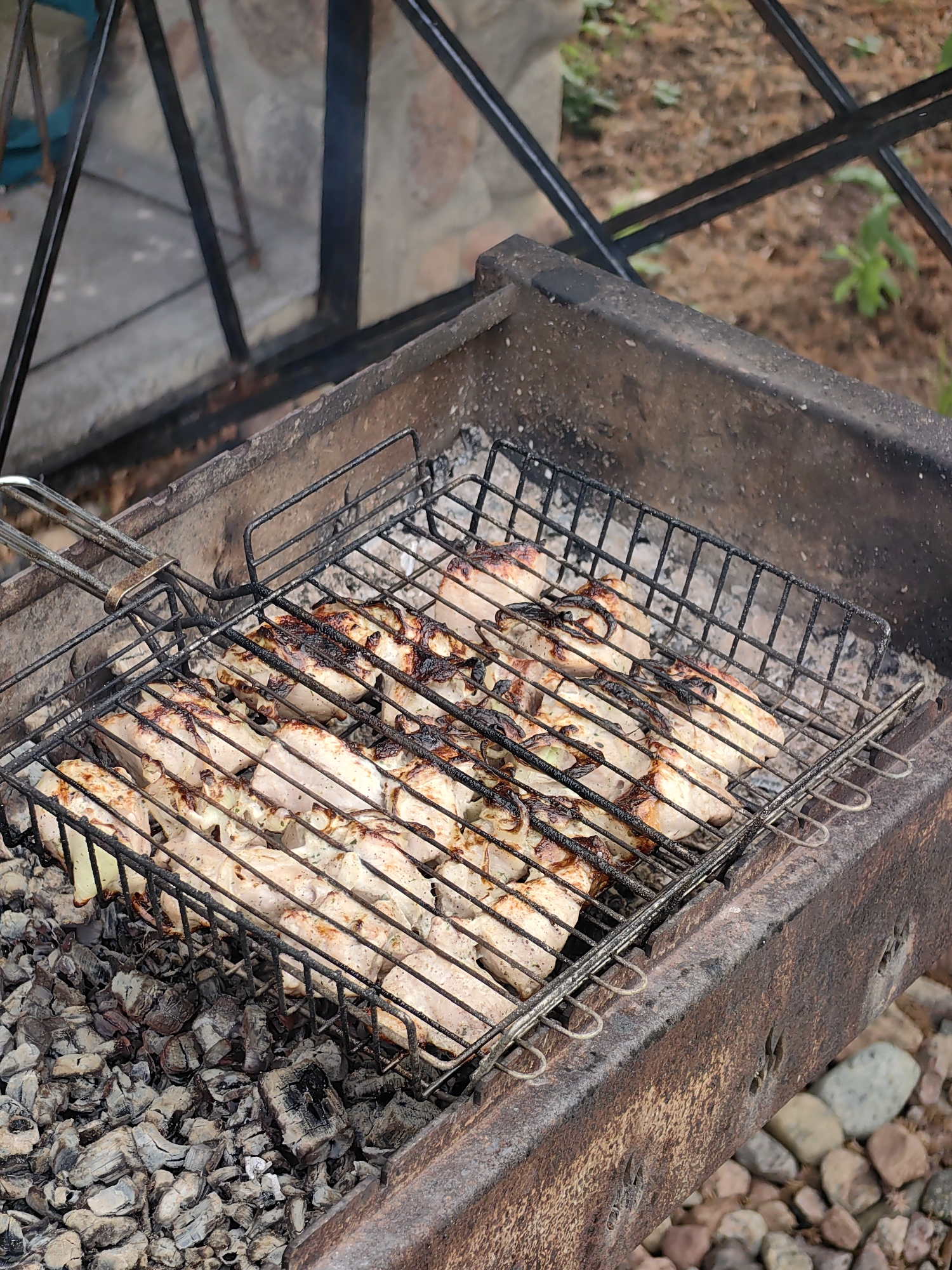 In Myadzyel, Belarus, a close-up, outdoor shot captures raw meat cooking on a barbecue grill. The grill, constructed from weathered metal, rests on a bed of hot charcoal, some pieces of which are still smoldering. The meat, appearing to be chicken pieces, is arranged in rows on the grill grate, some exhibiting browning and charring from the heat. The background shows a blurred outdoor environment with a stone structure and foliage. The image is likely taken during the day due to the lighting.