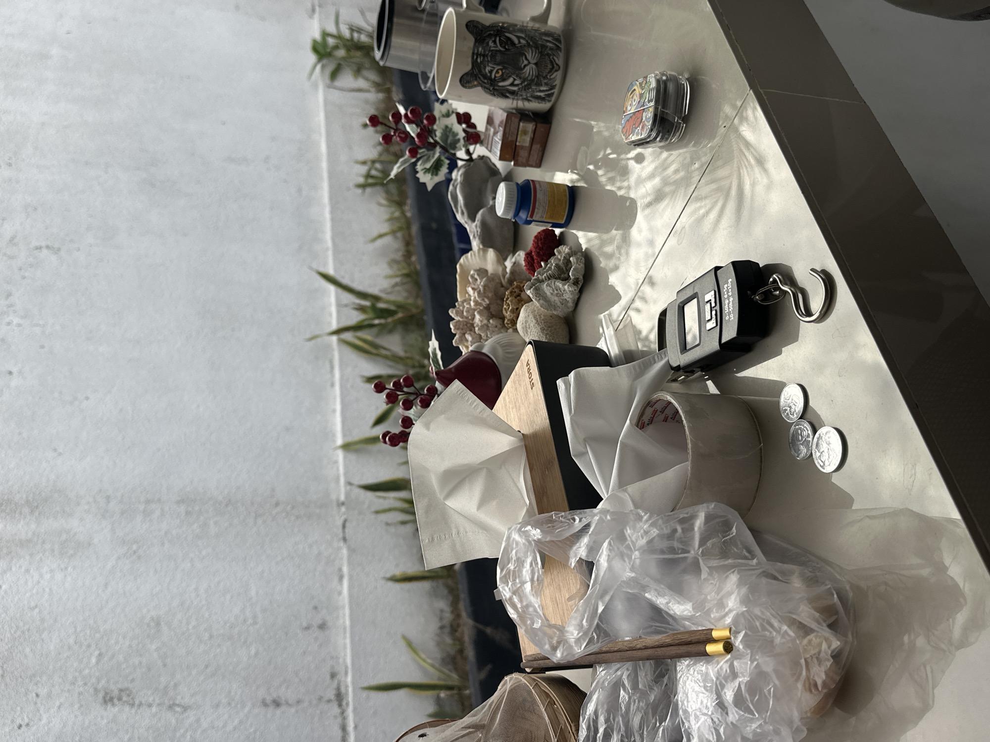 The image shows a cluttered surface with various items. There is a mug with a tiger design, a tissue box, a roll of tape, plastic bags, chopsticks, coins, a scale, and some decorative items, including coral and red berries. The background shows a wall with greenery. The location is North Kuta, Indonesia.