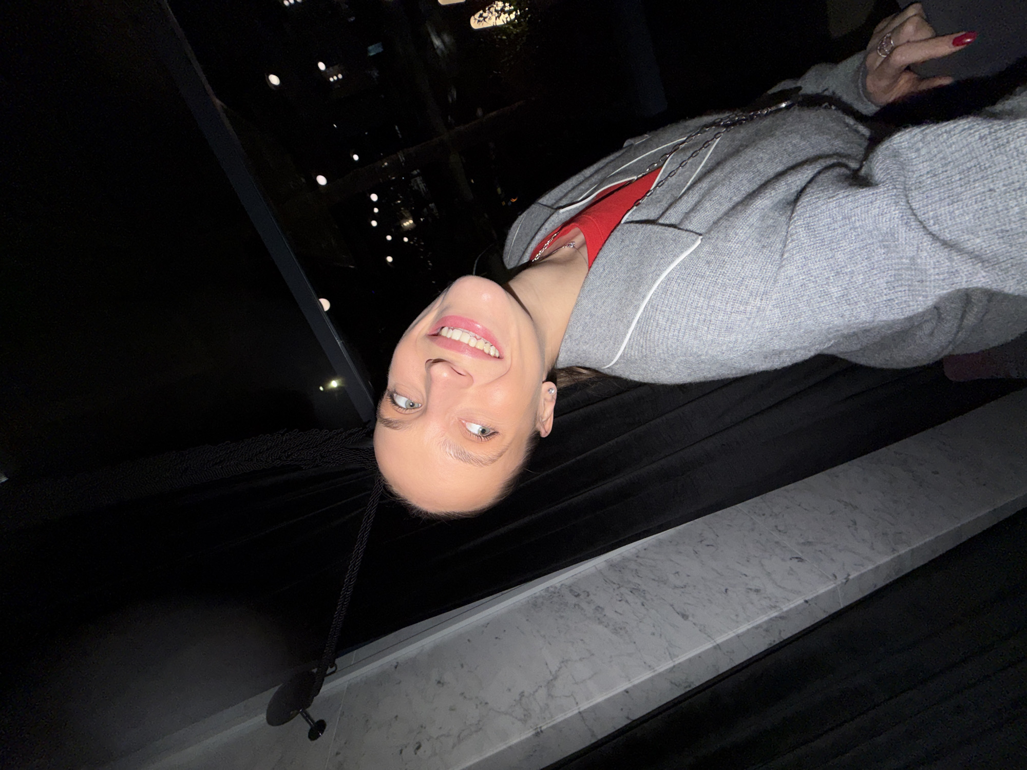 A woman is smiling and giving the middle finger in a selfie-style photograph taken at night in what appears to be a building in Budva, Montenegro. The background shows city lights at night, possibly seen through a window. The woman is wearing a gray sweater over a red shirt, and a silver chain is visible. She is standing next to a white marble-style ledge. The environment is dark, highlighting the interior lighting and the city lights outside.
