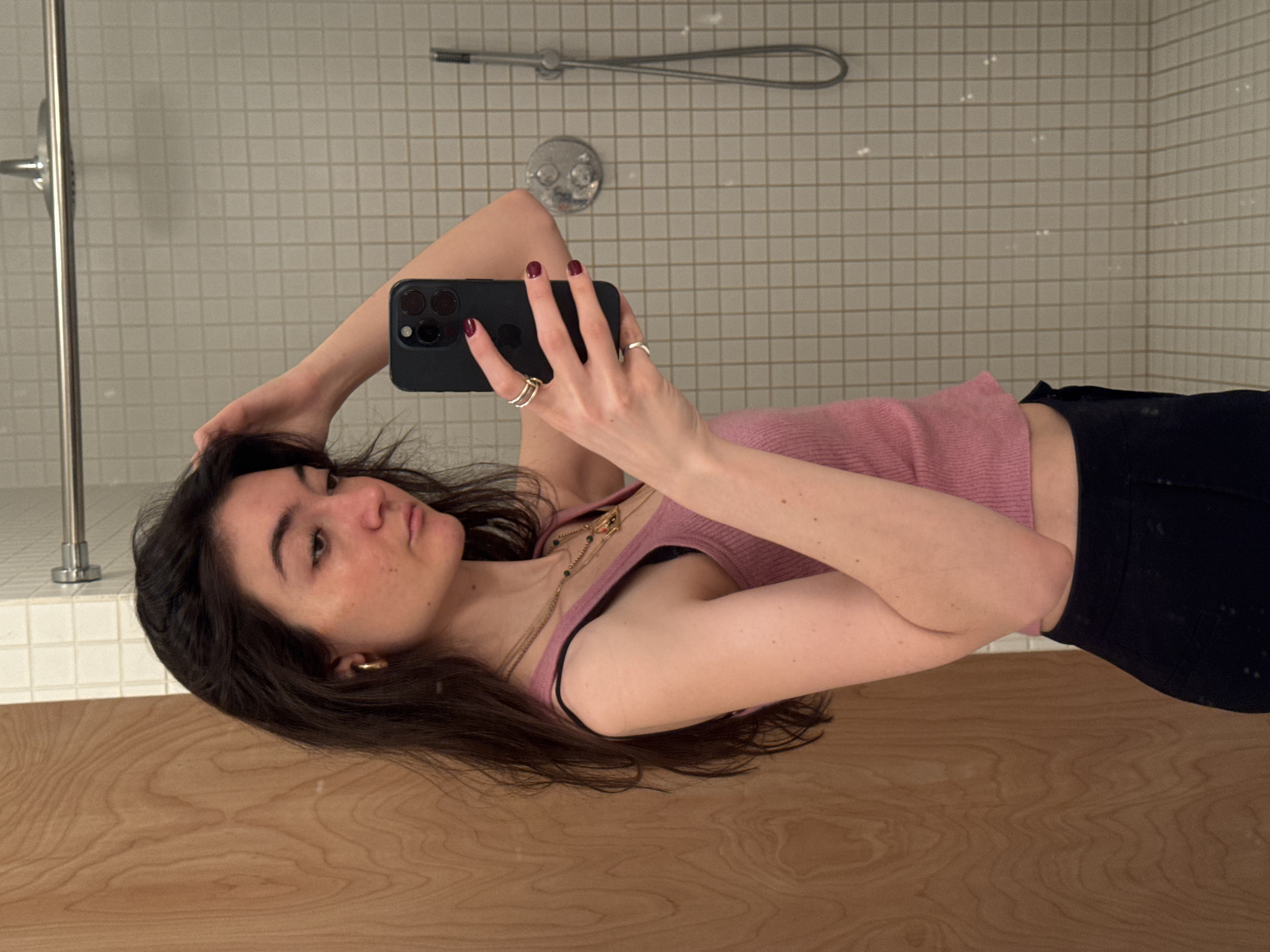The image displays a young woman taking a mirror selfie, with the photograph oriented sideways (rotated 90 degrees clockwise). The woman is positioned horizontally across the frame, with her head towards the left and her body extending to the right.
She has long, dark hair that frames her face and flows over her shoulders. Her skin is fair, and she has a calm expression, looking directly into the camera with slightly pouted lips. She is wearing a light pink, ribbed tank top, and a black bra strap is visible on her left shoulder. Around her neck, she wears a delicate, layered gold-colored necklace with small pendants. A simple gold hoop earring adorns her visible left ear.
Her right hand is raised, holding a black smartphone (identifiable as an iPhone Pro model due to the distinct three-lens camera module) towards her face to capture the reflection. Her fingernails are painted a dark red or burgundy color. On her right hand, she wears several rings: two thin, gold-colored bands on her ring finger and a single silver-colored band on her middle finger. Her left hand is placed casually on the back of her head, with her fingers intertwined in her dark hair. The lower part of her body shows she is wearing dark-colored pants or shorts.
The background, reflected in the mirror, reveals an indoor setting, likely a bathroom. The upper portion of the background (behind her upper body) is a wall covered with small, light-colored square tiles and darker grout lines. Mounted on this tiled wall are chrome bathroom fixtures, including a shower head with a flexible hose and a central faucet with two circular control knobs. The lower portion of the background (below her head and torso) consists of a smooth, light-colored wooden surface, possibly a countertop or vanity. This wooden surface provides a reflection of her hair and parts of her clothing.
The overall scene suggests a casual, personal moment captured indoors. There is no visible text in the image.