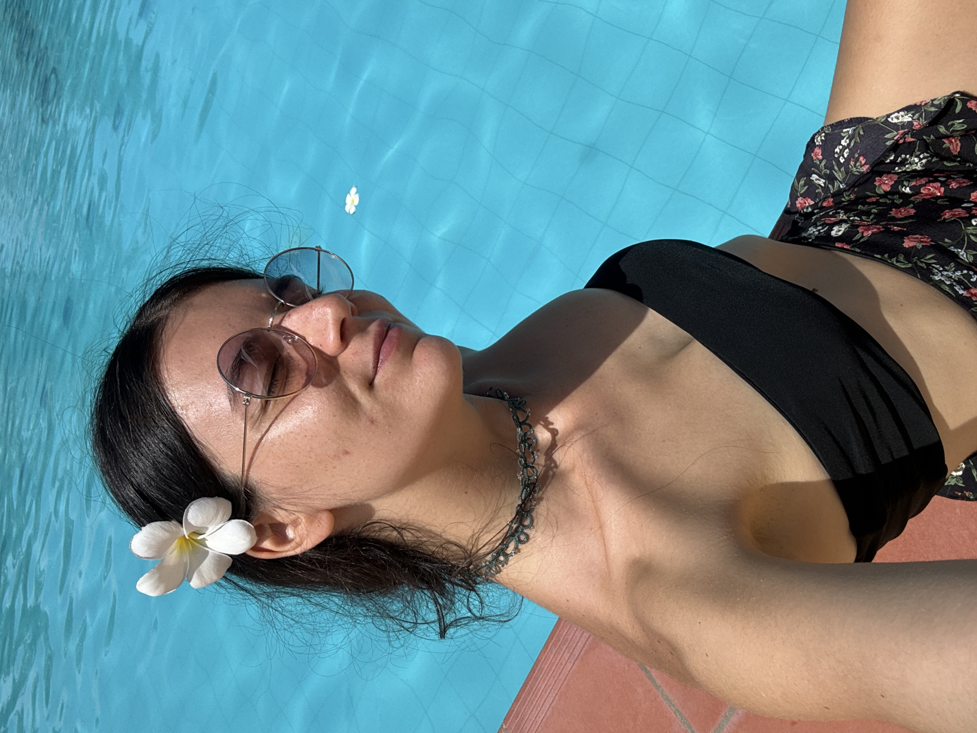 A female individual is depicted in an outdoor setting at the edge of a swimming pool in Phu Thuy Ward, Vietnam. She has dark hair and is wearing round, wire-rimmed sunglasses with light-colored lenses. A white plumeria flower is placed behind her left ear. Her attire consists of a black strapless top, a dark choker necklace with a patterned design, and a dark garment featuring a floral pattern of red and white flowers. Her eyes are closed or squinting, indicating direct exposure to sunlight. Her left shoulder and arm are positioned in the foreground. The background features a bright blue swimming pool with a visible tiled grid pattern on its bottom. A single white plumeria flower floats on the water's surface. The pool deck, composed of terracotta-colored tiles, is visible along the lower-left edge of the frame. The scene is illuminated by strong sunlight, casting shadows.