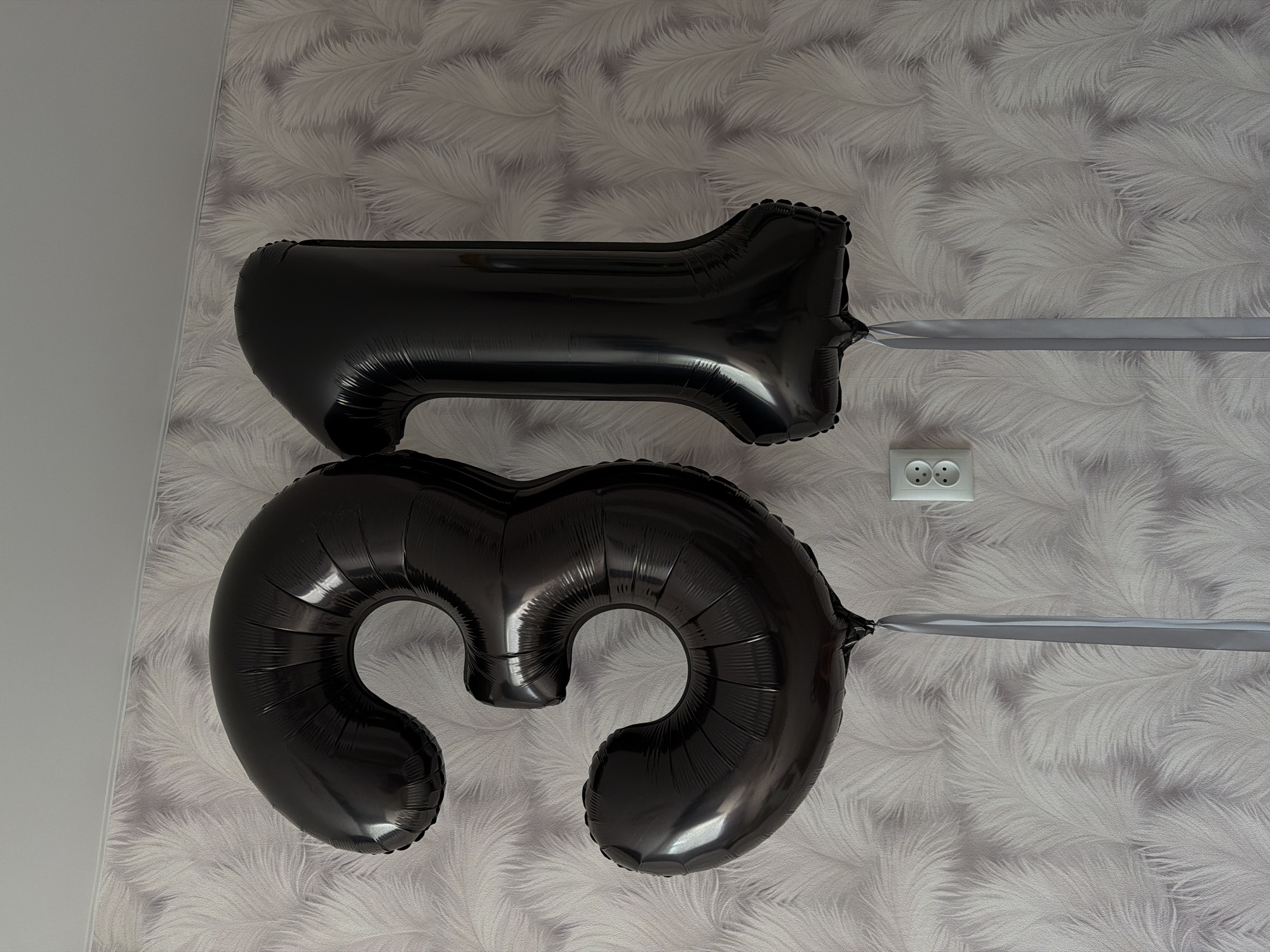 The image shows two inflated black foil balloons shaped like the numbers "1" and "3" against a wall covered in a patterned wallpaper. The balloons are attached with grey ribbons. An electrical outlet is visible on the wall to the right of the "1" balloon. The image was taken indoors and is oriented in portrait format. The image does not provide enough information to determine the precise location of the picture.