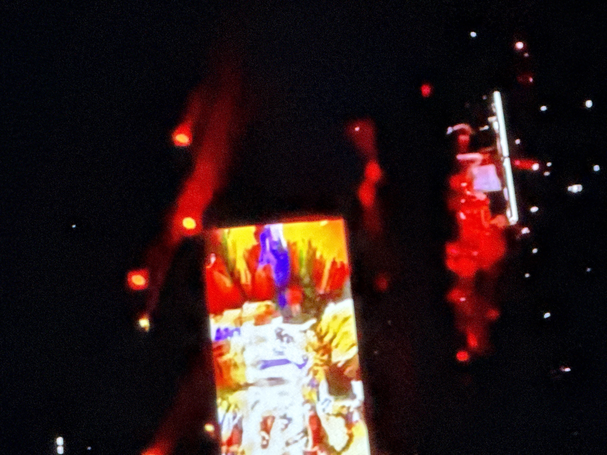 The image depicts a dark, high-contrast scene, likely from a concert or live event in a low-light setting. The central focus is a brightly illuminated, vertically oriented rectangular screen displaying vibrant and abstract imagery. The screen exhibits a chaotic mix of colors, predominantly vivid yellow, bright red, deep blue, and white, forming indistinct figures or graphical patterns. A distinct blue shape in the upper part of the screen could be a stylized logo or text, while white and yellow elements below suggest dynamic figures or designs.
The background is largely dark black, punctuated by numerous small, bright white dots resembling distant lights or reflections. Several larger, blurry red and orange light sources appear as streaks or glows, suggesting dynamic stage lighting, pyrotechnics, or motion blur from lights held by individuals. A prominent vertical white line, possibly a light fixture or structural element, is visible towards the right side of the frame, amidst some of the red blurs and white dots. The scene conveys an atmosphere of energy and visual spectacle. Due to blurriness and pixelation, no specific text is clearly legible on the screen or elsewhere. The time of day is definitely night, or the event is taking place in a darkened indoor venue.