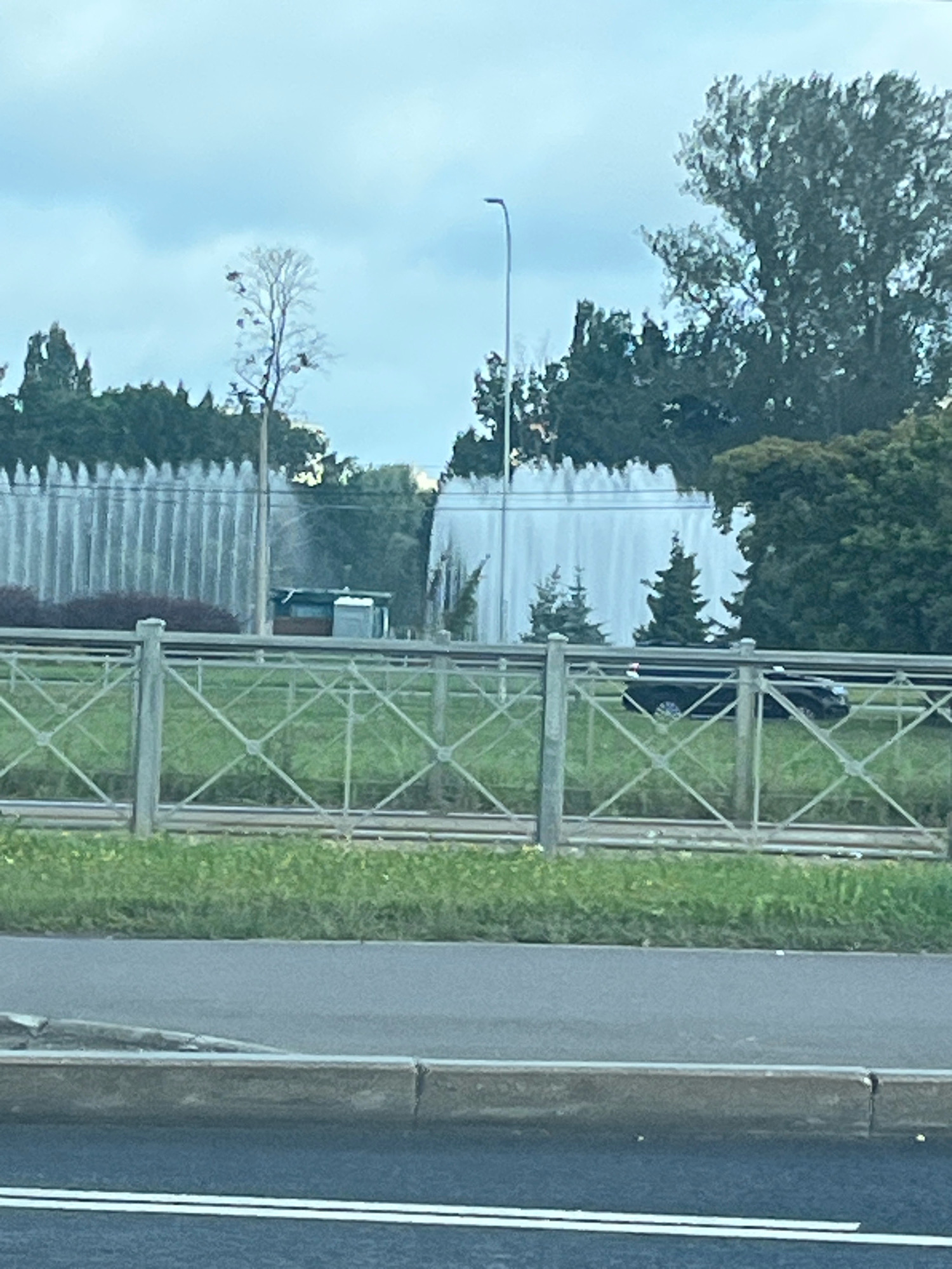 An outdoor, slightly overcast, medium-shot image captures a scene in Saint Petersburg, Russia. In the foreground, a gray curb with two white horizontal lines marks the edge of a road. Behind the curb is a sidewalk, and then a metal diamond-patterned fence. Beyond the fence, a strip of green grass is visible, with a train track running parallel to the fence. In the mid-ground, a large white structure, possibly a fountain or a misting system, is active, creating a vertical spray of water. A small, dark-colored structure, possibly a portable toilet or utility building, is partially visible behind the spray. Further back, evergreen trees and other foliage form a dense backdrop. A dark-colored car is driving on a road behind the fence, partially obscured by the water spray. In the far background, taller trees and the sky are visible.