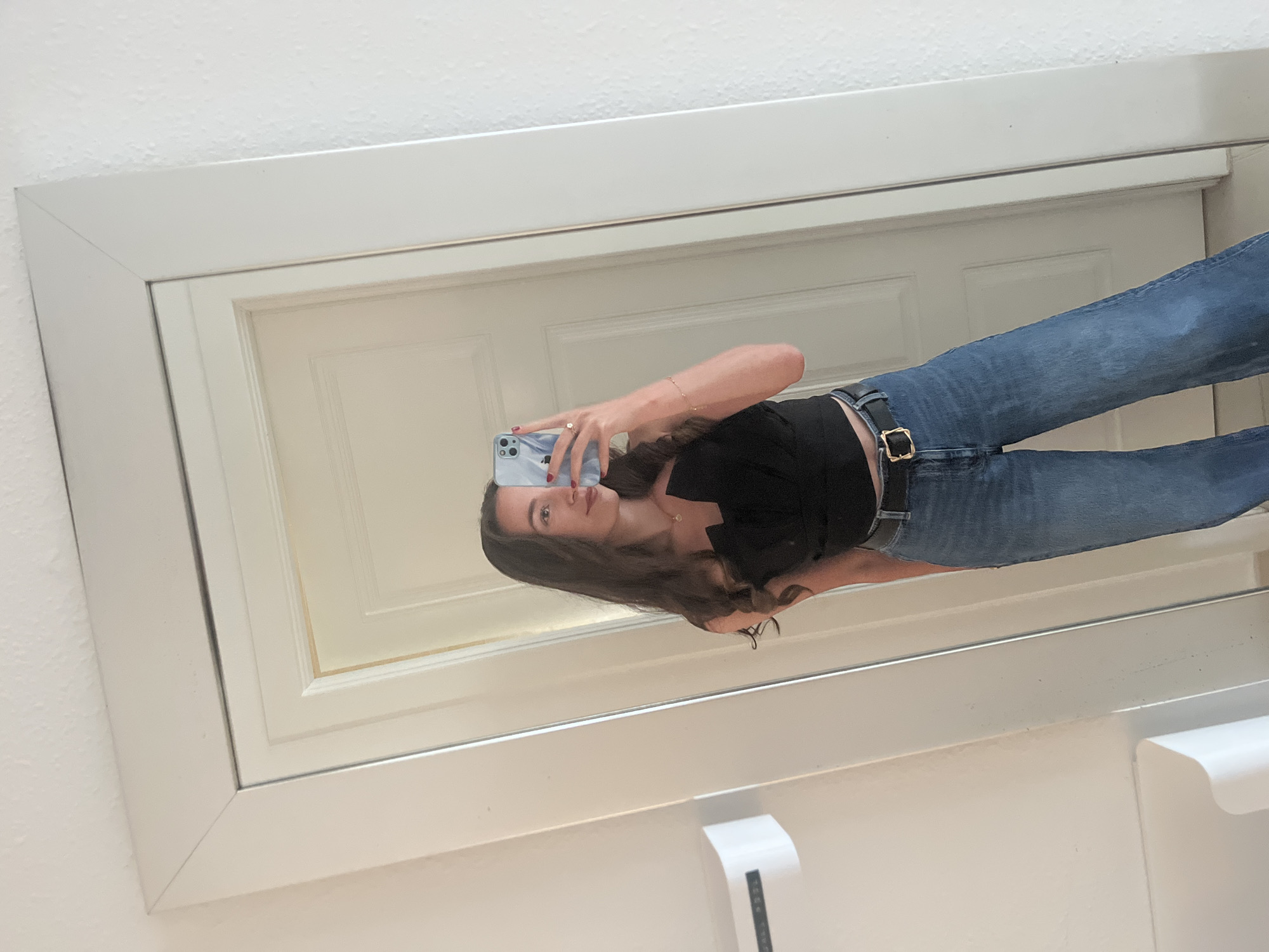 The image shows a person taking a selfie in front of a mirror. The person is wearing a black top and blue jeans with a black belt. The mirror has a white frame. A white door and a hand soap dispenser can be seen in the background. The location appears to be indoors, possibly a bathroom or dressing room. There is no specific information to determine that the location is El Poble Nou de Benitatxell / Benitachell, Spain.