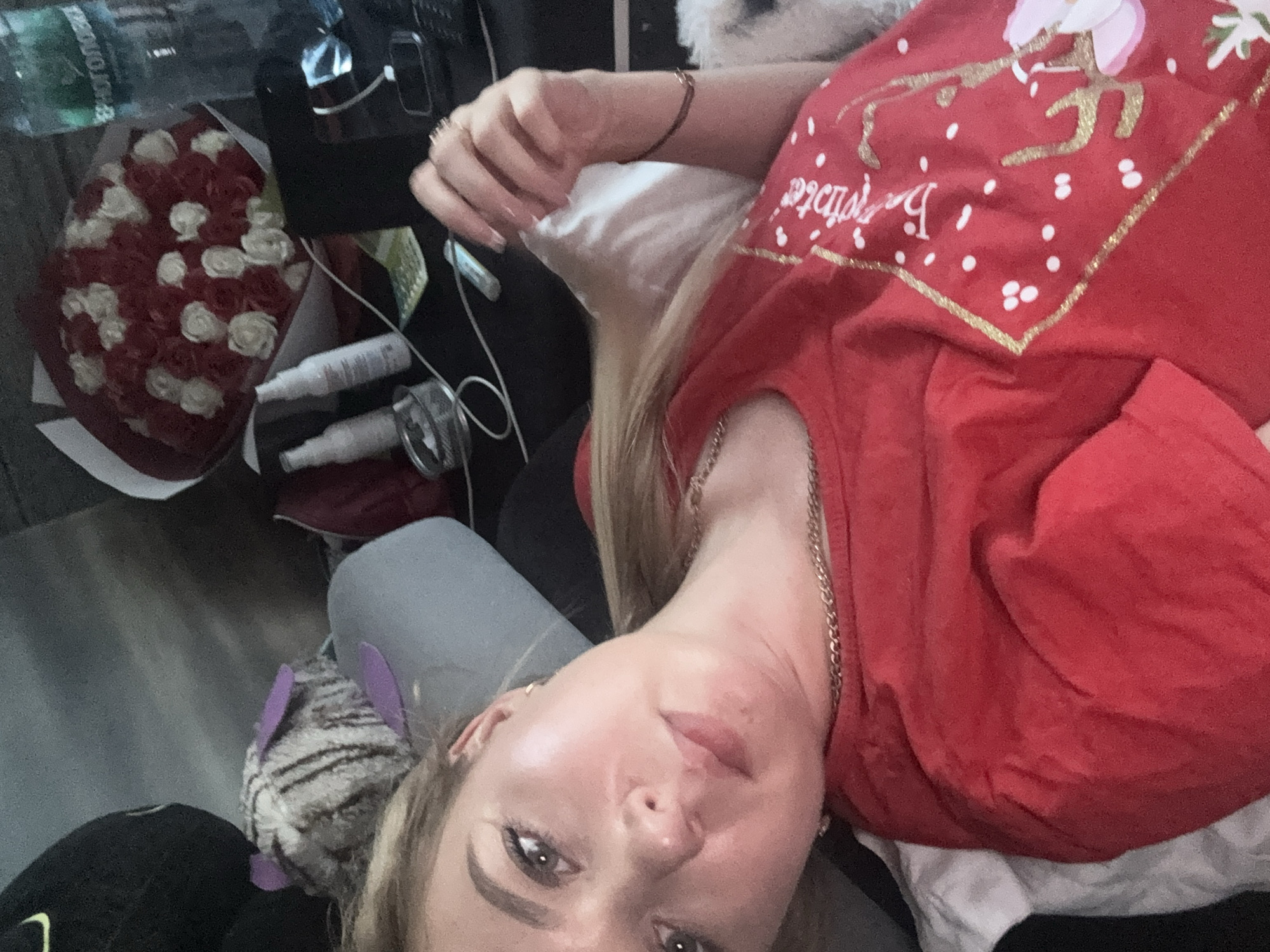 The media file shows a selfie of a woman lying down, with her face close to the camera. She has blonde hair and is wearing a red shirt with a gold and white design. She is wearing a necklace. To the left of the woman is a bouquet of red and white roses. There are also several bottles and some electronic devices in the background. The woman appears to be indoors, possibly in a bedroom or living room. The lighting suggests it could be daytime. The overall mood seems relaxed. The text visible on the shirt appears to be "Be Festive" in cursive.