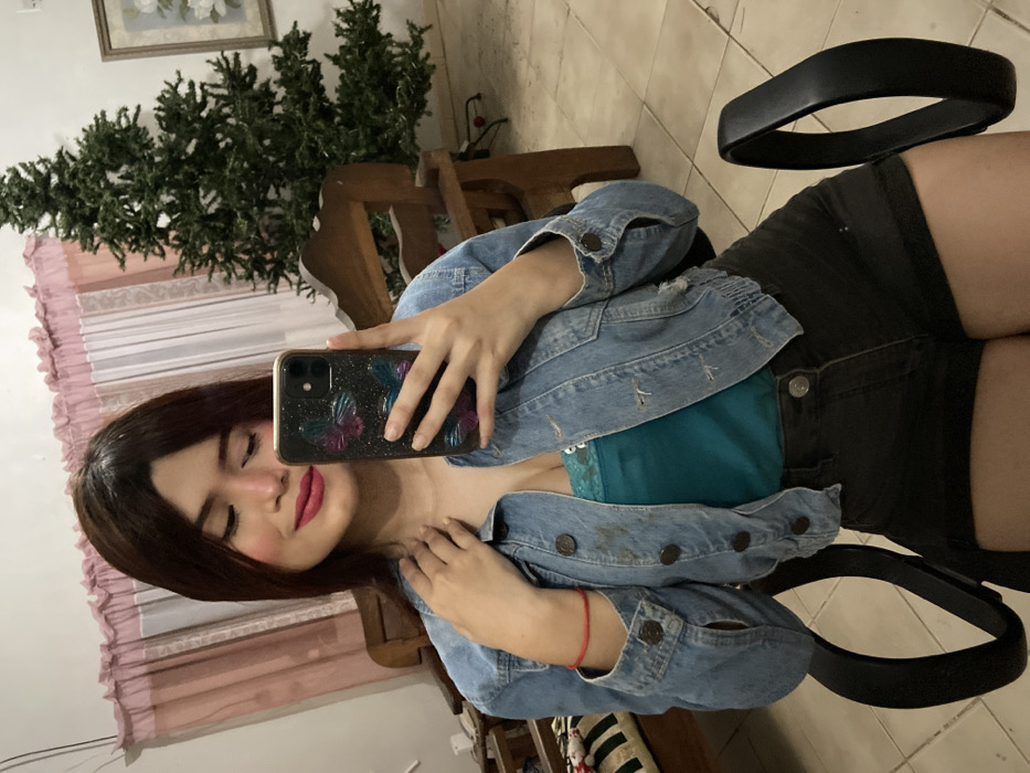 A woman is seated, facing left, in a chair with black armrests. She's taking a selfie with her phone, which has a case adorned with butterflies. She's wearing a blue denim jacket over a green top and black shorts. Her hair is brown. Behind her, a Christmas tree stands near a window with pink curtains. A wooden chair and other furniture are also visible in the background.