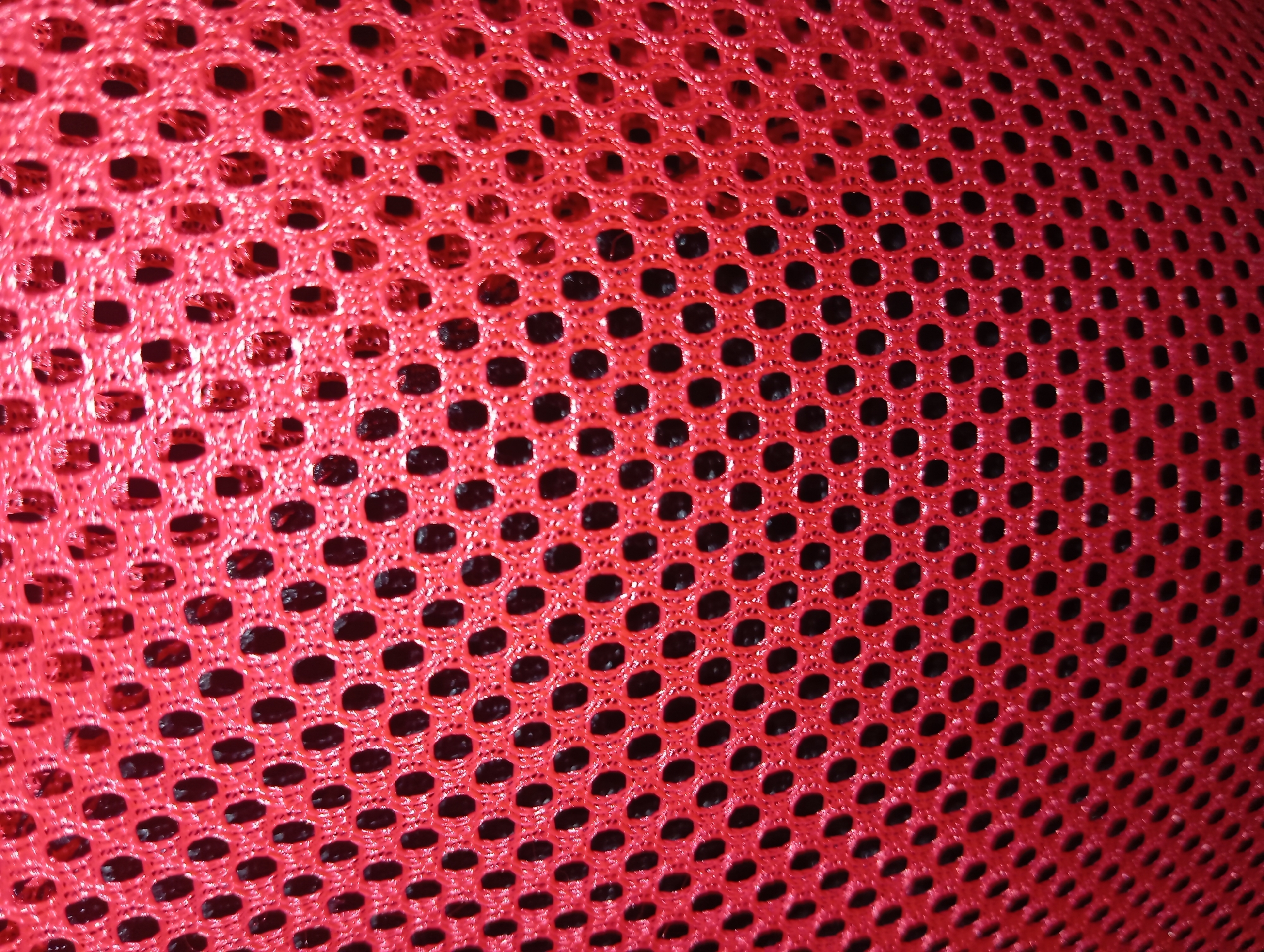The image shows a close-up, abstract view of a red, mesh-like fabric with hexagonal or circular holes. The texture of the fabric is visible, with a slightly shiny and woven appearance. The holes reveal a dark, possibly black, background. The lighting creates highlights on the red material, giving it a three-dimensional quality. There is no discernible context within the image that would place it in Rejosari, Indonesia, or any other specific location, nor are there any people, subjects, actions, or interactions depicted. The image is purely a textural study of the fabric.