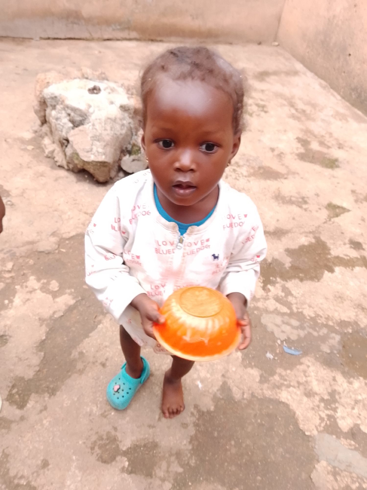 A young child stands outdoors on a concrete ground, holding an orange bowl. The child is wearing a white long-sleeved shirt with blue trim and a pink design on one shoe. A large rock formation is visible behind the child. The background appears to be a wall. The lighting suggests it is daytime. The location is unknown.