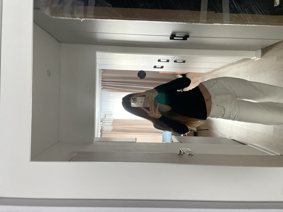 A mirror selfie captures an individual standing in an interior space identified as Khimki, Russia. The person, with long brown hair, holds a light-colored smartphone horizontally, partially obscuring their face, to photograph their reflection. They are dressed in a black long-sleeved top, exposing the midriff, and white wide-leg trousers. The reflected background reveals a residential interior hallway with light-colored walls and wooden flooring extending into an adjacent room. Visible in the reflection are a doorframe with a silver handle to the left, a segment of a bed with a blue duvet, a crystal-strand chandelier, and light peach-colored curtains. To the right, a sequence of white built-in wardrobes with black handles is visible, alongside a large, clear-plastic-wrapped object and a dark circular wall-mounted item. A green lens flare is present on the smartphone's reflective surface.