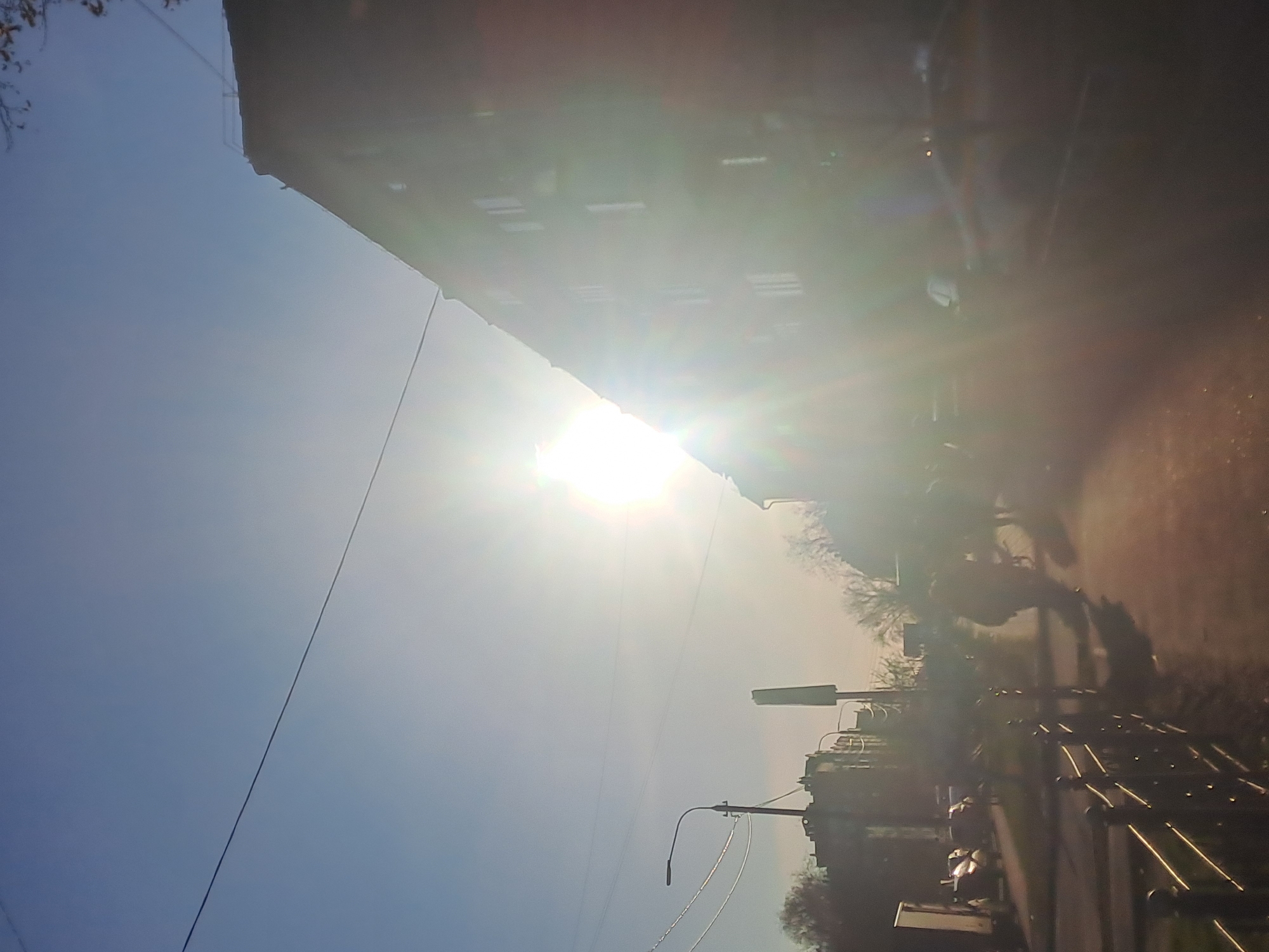 The image shows a street scene in Elektrostal, Russia, on a sunny day. The sun is high in the sky, causing glare. A multi-story building stands on the left side of the frame. A power line is visible against the blue sky. A sidewalk runs along the right side of the image, lined with a metal railing. Further down the street, there are street lights, trees, and other buildings. Pedestrians and parked cars are visible in the background.