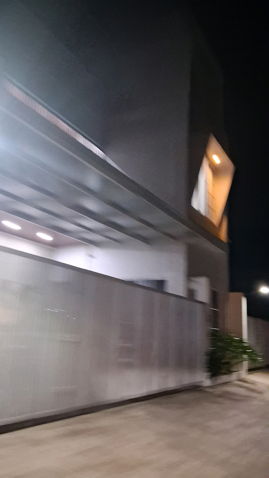 The image shows a blurry, nighttime view of a modern building exterior and surrounding area. In the upper left, a section of the building features a horizontal canopy supported by beams. Below the canopy, there are multiple recessed circular lights on a ceiling. A tall, light-colored wall or fence runs horizontally across the middle of the frame, with a darker strip along its top edge. To the right of the wall, a small area of green foliage is visible. In the upper right portion of the image, an angular architectural feature of the building is illuminated from within, casting a warm glow. The overall scene is dimly lit by artificial light sources, with some motion blur suggesting movement. The image is set in the city of Madurejo, Indonesia.