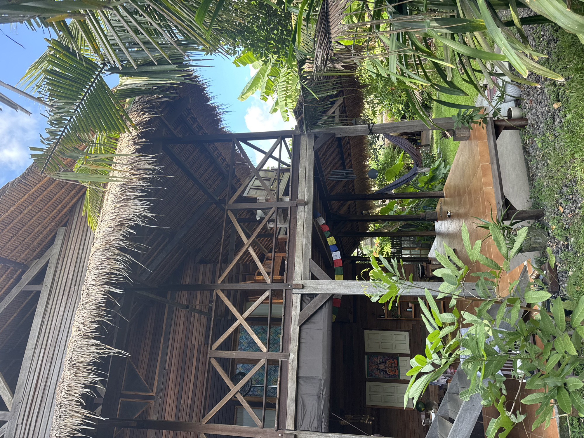 This media file captures a serene, rustic scene in Kediri, Indonesia, featuring a multi-level wooden structure with a prominent thatched roof. The image, though rotated, depicts a tall, open-sided building constructed from dark timber planks and beams, featuring intricate cross-bracing details. Its architecture, with exposed wooden framework and a natural fiber roof, suggests a traditional or eco-friendly tropical design.
The building is enveloped by lush tropical vegetation. Large palm fronds frame the upper edges, while banana leaves and various other broad-leafed plants contribute to a dense, jungle-like atmosphere. Patches of clear blue sky with soft white clouds are visible through the canopy and between the building's structural elements.
Notable details within the scene include a relaxed setting. A hammock is strung on the right side of the structure, inviting repose. Colorful prayer flags, strung horizontally, add a vibrant touch. On the lower level of the building, closed wooden window shutters and a framed artwork or tapestry adorn a wall. A ceiling fan can be glimpsed through an open window space. Adjacent to the building, a tiled or paved outdoor area suggests a patio or veranda, with glimpses of green grass and pebbled ground nearby. Wooden stairs are also visible, leading to different levels of the structure. A black, cylindrical item, possibly a rolled mat or sleeping bag, rests against a wooden post.
There are no people visible, and no specific activity or event is taking place, contributing to the tranquil ambiance. The natural lighting suggests it is daytime, likely mid-morning or early afternoon, with fair weather. No visible text is present in the image. The overall impression is one of a peaceful retreat integrated into a vibrant natural environment.