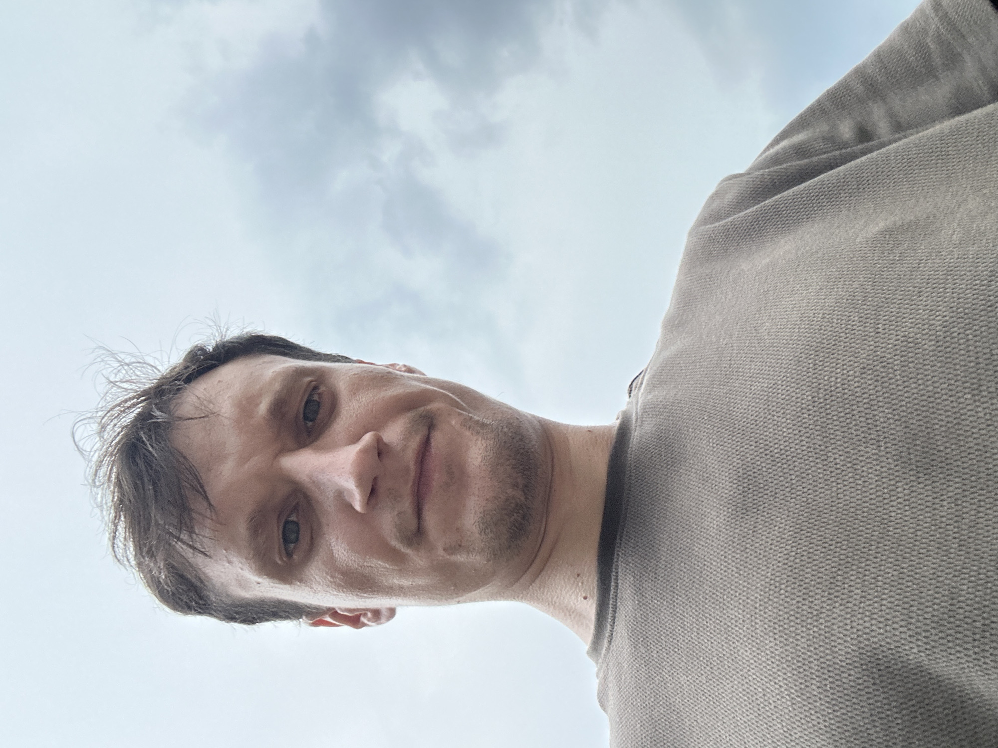 A low-angle, close-up selfie of a man's face and chest is shown against a cloudy sky. The man has short, messy brown hair, light skin, and appears to be in his late 20s or early 30s. He has a slight stubble on his chin and upper lip. He is wearing a plain, light brown t-shirt with a textured fabric. The background is predominantly a light gray, overcast sky with some darker clouds. The image was taken outdoors, likely in the city of Kamianets-Podilskyi, Ukraine, though no specific landmarks are visible. The man is looking directly at the camera with a neutral expression.