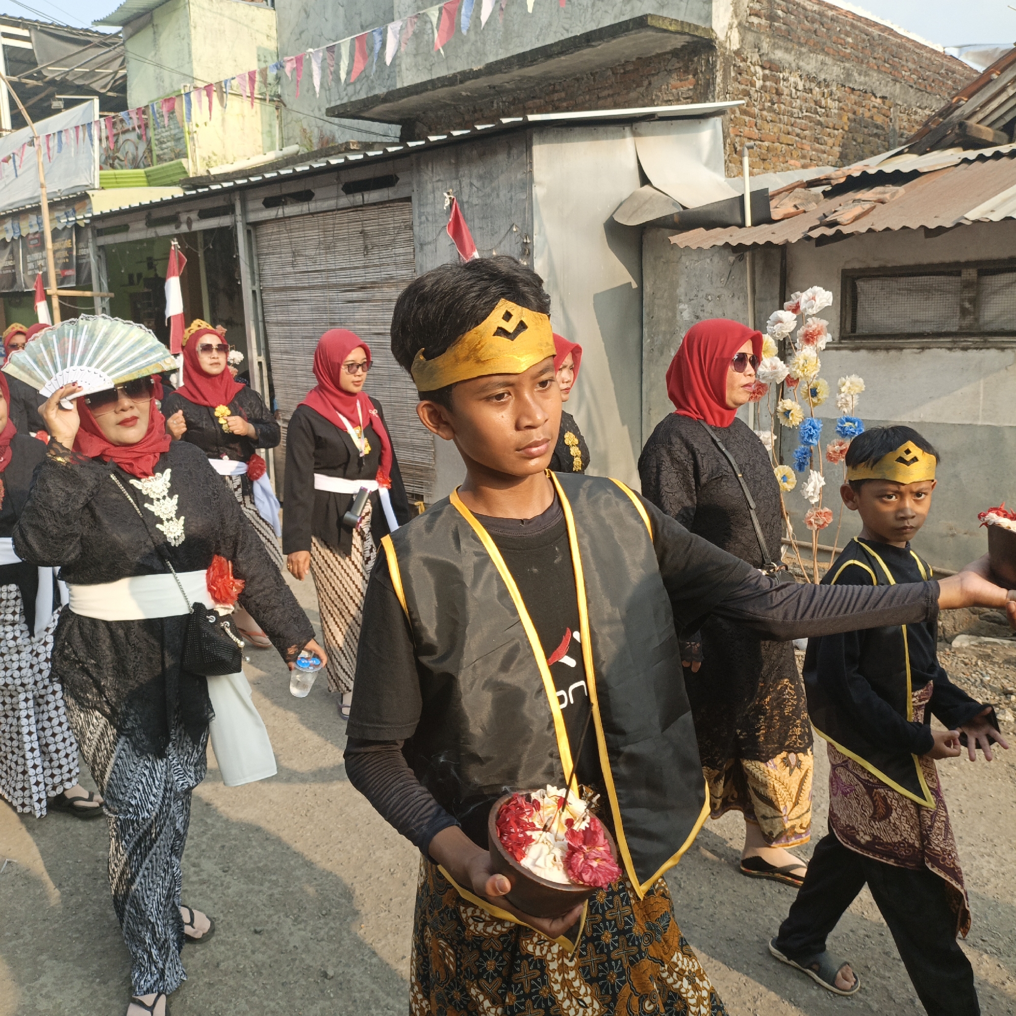 In Tanahbaya, Indonesia, during what appears to be a cultural parade or ceremony, a group of individuals, predominantly women wearing red hijabs and traditional black lace tops with patterned sarongs, are walking down a street. One woman in the foreground holds a large, decorative fan. In the center of the image, a young boy, adorned with a golden crown and a dark vest trimmed with yellow, carries a bowl filled with colorful petals and an incense stick. Another young boy in a similar outfit is visible to his right. The street is lined with buildings, some appearing older with corrugated metal roofs, and festive banners are strung overhead. The ground is paved with asphalt, and debris is visible on the sides. The lighting suggests it is daytime, possibly late afternoon.