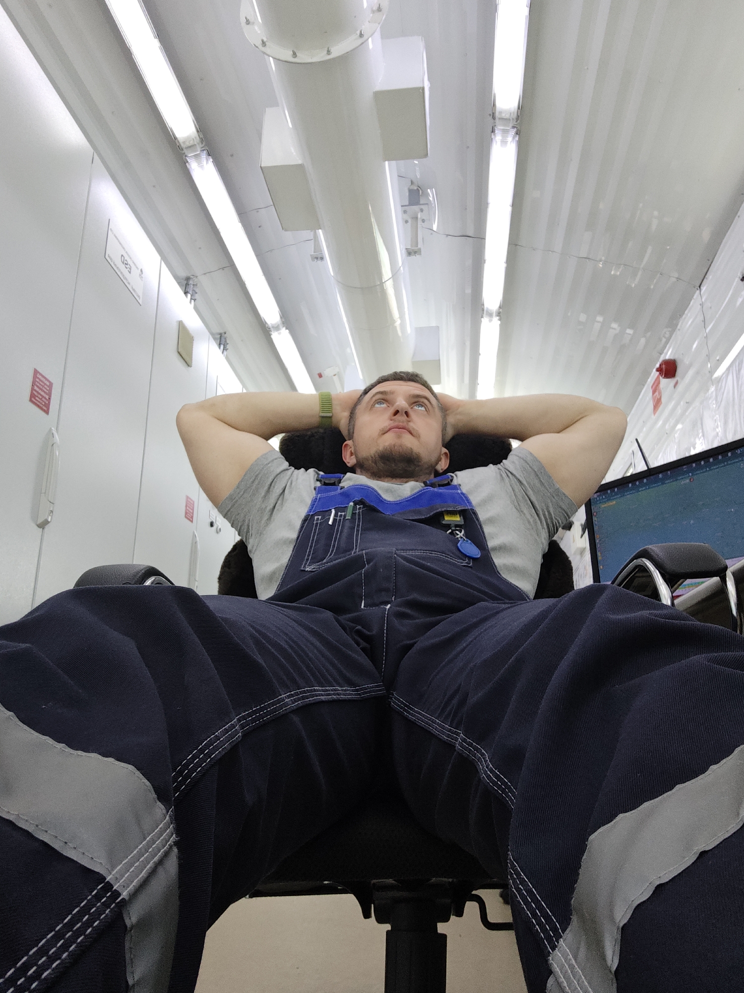 A low-angle shot depicts a man seated in an office chair, his hands clasped behind his head, looking upwards. He is wearing a grey t-shirt and blue overalls with reflective stripes on the legs. The office features a white ceiling with fluorescent lighting and cylindrical pipes. A computer monitor is visible on the right side of the frame, along with wall-mounted equipment. The location is unknown.