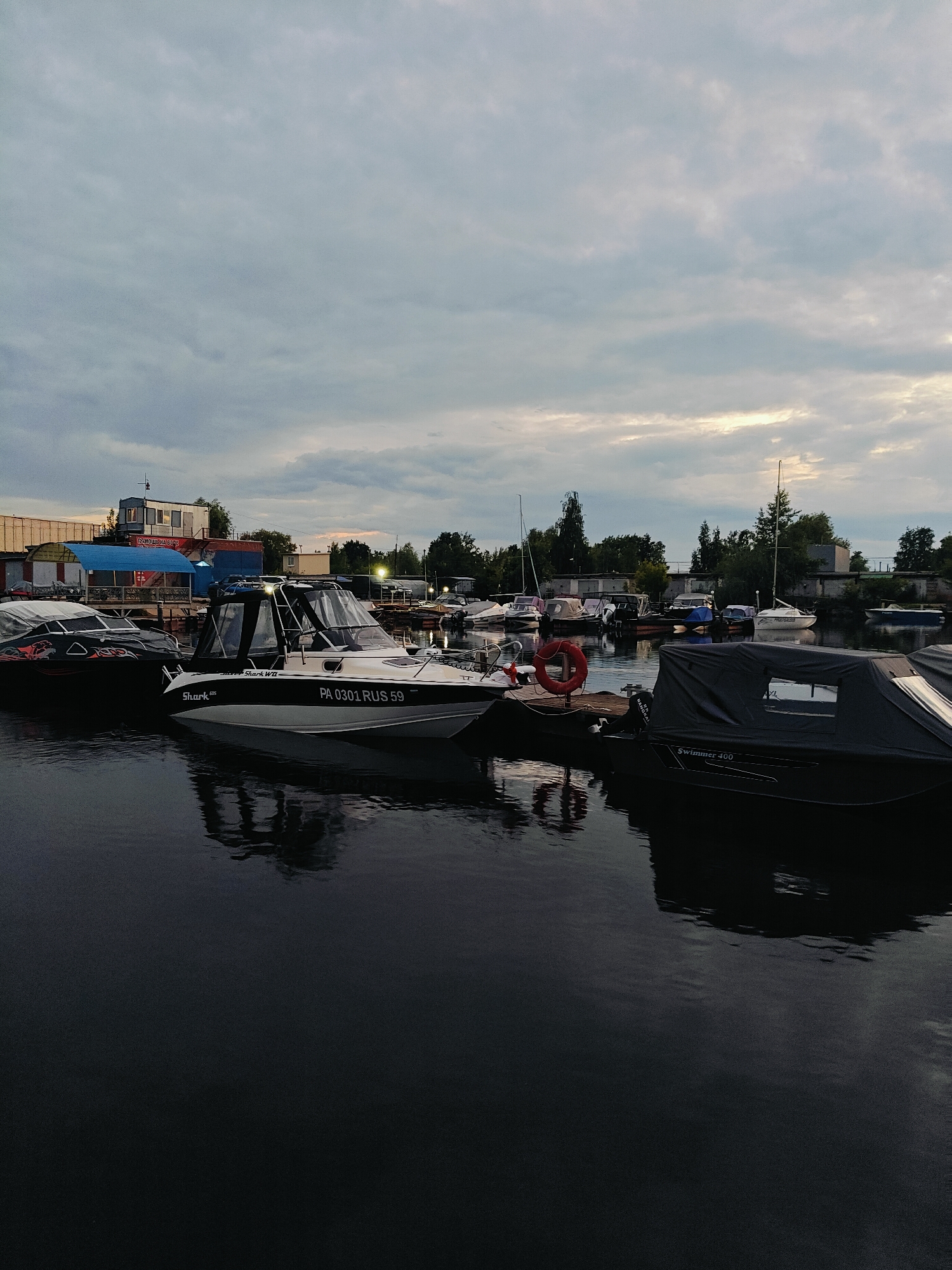 The image depicts a twilight scene at a boat marina, likely in the city of Chaikovsky, Russia, given the license plate "RUS 59" visible on a boat. Several boats are moored along a wooden dock. In the foreground, a dark-covered boat is visible, with the text "Swimmer 400" on its side. Next to it, a white and black boat with a cabin is docked, displaying the license plate "PA 0301 RUS 59" and the name "Shark WD." To the left, another black boat with a red and black graphic design is partially covered. In the background, several other boats are moored, and buildings and trees line the shore. The sky is overcast with dramatic cloud formations. The water is calm and reflects the boats and the sky. There are no people visible in the image. The scene suggests a quiet evening at the marina.
