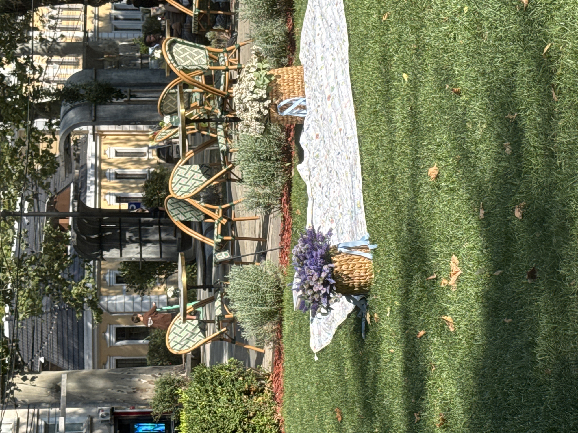 The image depicts an outdoor seating area of a cafe or restaurant, possibly located in the city of Chișinău, Moldova. In the foreground, a picnic blanket is spread on a well-maintained green lawn. Two wicker baskets filled with lavender and white flowers are placed on the blanket. In the mid-ground, several tables and chairs with a green and white patterned upholstery are arranged on a paved area, suggesting an outdoor dining setup. Behind the tables, lush green bushes and trees provide shade. In the background, architectural structures, possibly buildings or ornate archways, are visible. The scene is illuminated by natural daylight, casting shadows on the grass. There are a few fallen leaves scattered on the lawn. The overall impression is one of a relaxed outdoor gathering space.