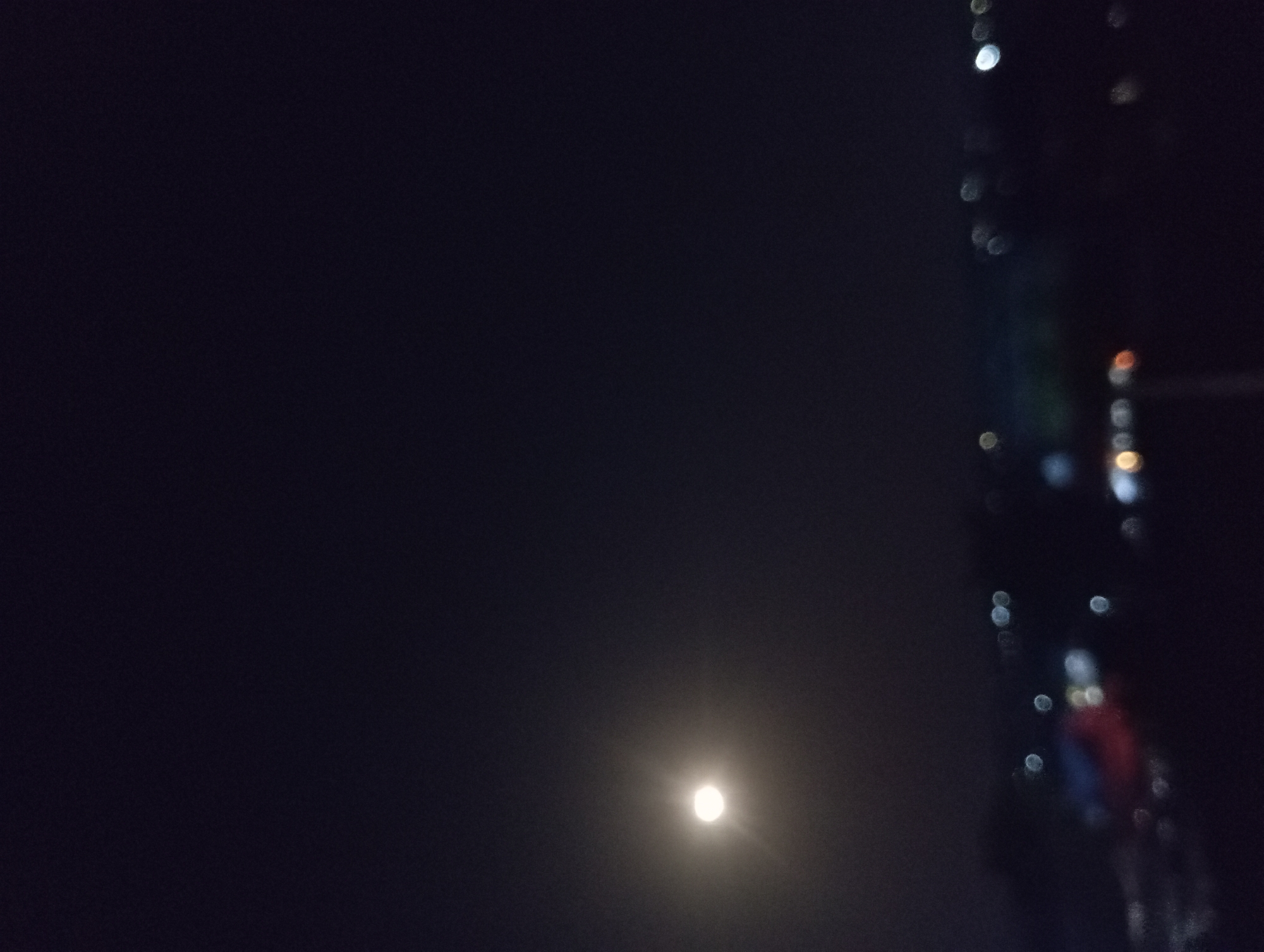 The image captures a tranquil nighttime scene, likely in Nkpor, Nigeria, featuring a celestial body and distant artificial lights. A bright, circular light source, resembling the moon, is prominently positioned in the lower-central part of the frame. It glows softly, casting a diffuse halo against the dark, featureless night sky, indicating either a hazy atmosphere or light cloud cover.
To the right, a vertical strip of numerous out-of-focus lights creates a bokeh effect. These blurred lights vary in intensity and color; some appear bright white or silver, while others emit warmer yellowish and orange tones. Closer inspection reveals hints of blue and red within these lights, suggesting diverse light sources from an urban environment, possibly illuminated windows of a tall building or streetlights lining a distant structure. The scene is static, devoid of any discernible human activity or specific objects other than the lights. The time of day is clearly night. No visible text is present. The overall mood is one of quiet solitude, contrasting the natural moonlight with the distant glow of human habitation.