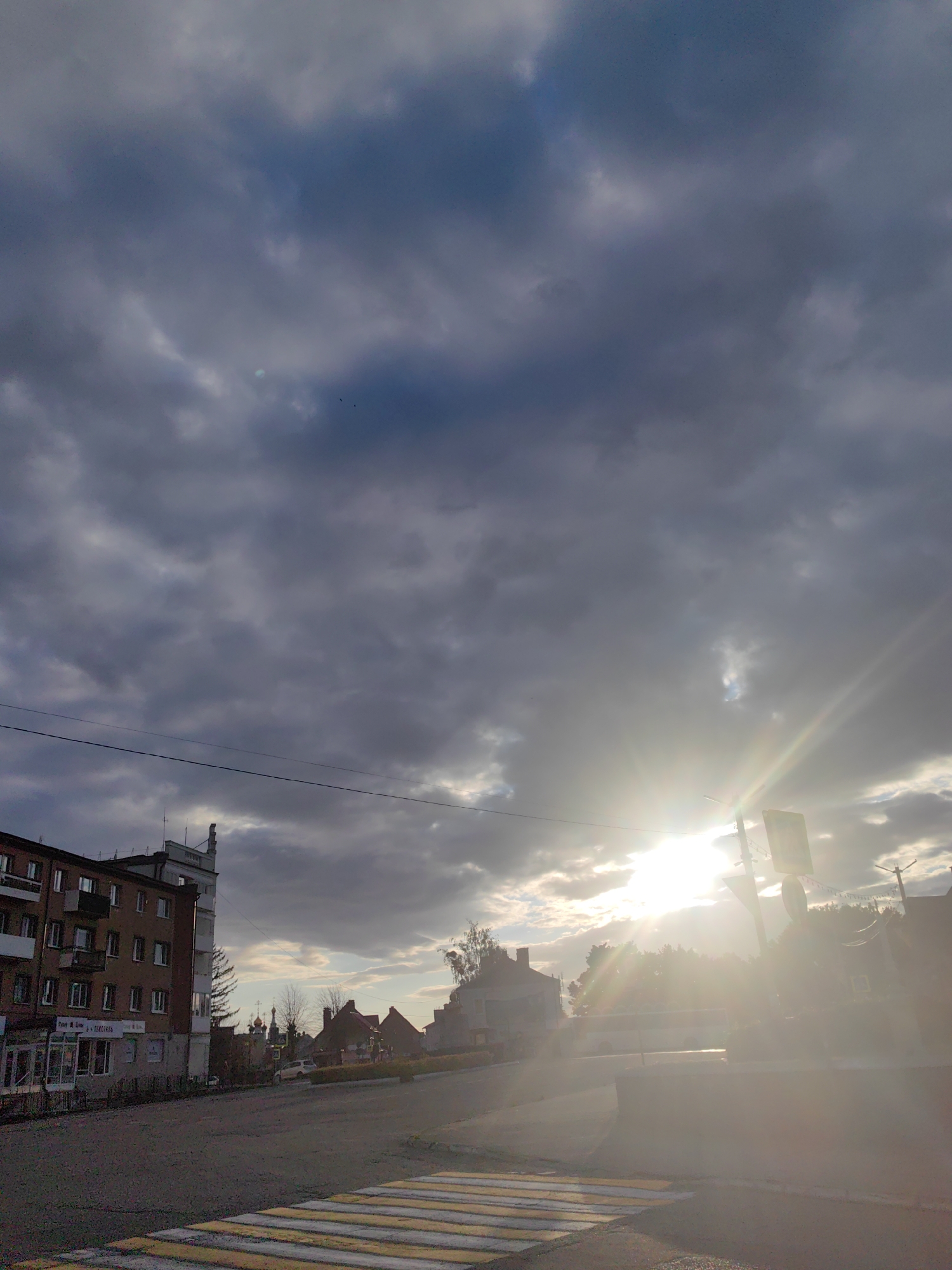 The image depicts a street scene in Bagrationovsk, Russia, under a dramatic sky. The foreground features a pedestrian crosswalk with yellow and white stripes. To the left, a multi-story brick apartment building with balconies and shops on the ground floor dominates the view. The shops have signage, with one clearly visible sign reading "TEKSTILNAYA" which translates to "Textile."
In the mid-ground, the street curves slightly, leading towards a cluster of buildings including what appears to be a church with golden domes. Trees with sparse foliage suggest autumn or early spring.
The sky is filled with heavy, dark clouds, but the sun is breaking through in the upper right quadrant, creating strong lens flare and illuminating the scene with a warm, ethereal glow. The contrast between the dark clouds and the bright sunlight creates a dynamic and somewhat moody atmosphere, hinting at a transition in weather. The time of day appears to be late afternoon or early evening, given the angle of the sun and the quality of light. The overall impression is one of a quiet, ordinary moment in a small Russian town, momentarily enhanced by the striking interplay of light and shadow in the sky. There are no people visible in the image.