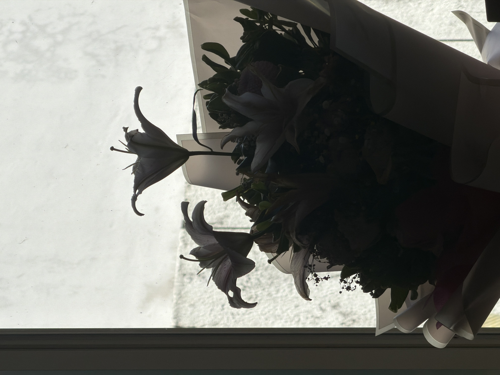 The image displays a strongly backlit silhouette of a flower bouquet, positioned indoors in front of a window. Two prominent lilies are visible at the top, their distinct petals, stamens, and pistils outlined against the bright background. Below these, a dense arrangement of other flowers and foliage forms a dark mass, partially enclosed by lighter-toned wrapping paper visible at the bottom and right. A dark vertical window frame is present on the far left. The outdoor background, viewed through the window, is uniformly bright and diffuse, indicating an overcast sky or reflective surface; faint specks and subtle textural variations are visible across this area. No visual information within the image provides specific indicators for the city of Kuta Selatan, Indonesia. No individuals are present.