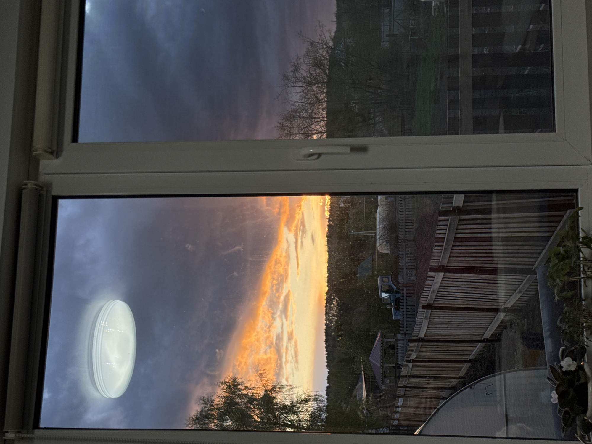 The image is taken from inside a building, looking out through a double-paned window. The top window shows a darkening sky over what appears to be vegetation, possibly trees and fields. The bottom window captures a dramatic sunset with orange and yellow clouds. Outside the bottom window, there is a view of a rural landscape, including a fence, greenery, and a greenhouse. The bottom window also has a reflection of an indoor light fixture. Krilovo, Russia, is not explicitly confirmed, but the rural setting suggests a location in a more remote area, consistent with Krilovo.
