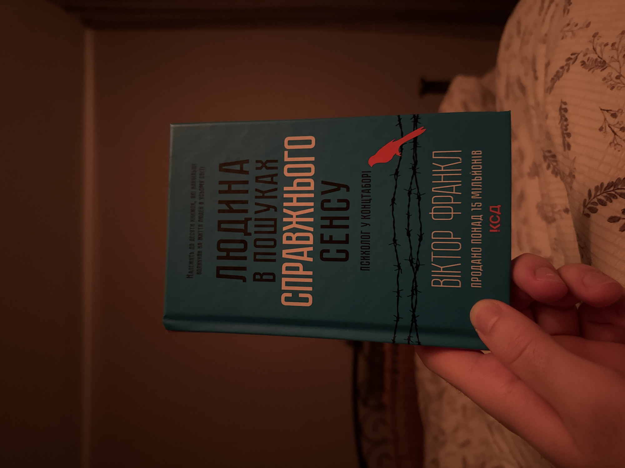 A person is holding the Ukrainian edition of Viktor Frankl's "Man's Search for Meaning." The cover features barbed wire imagery and a red bird. The text is in Ukrainian. The background is a dark, warm-toned setting.