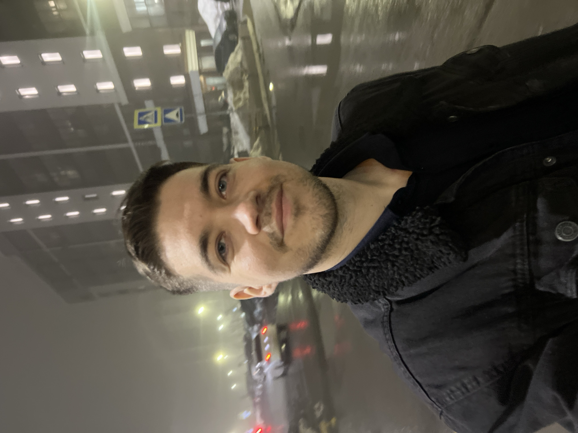 A male subject, appearing to be in his late 20s to early 30s, with short dark hair, light blue eyes, and a light beard, is centrally positioned, facing the camera with a faint smile. He wears a dark collared shirt and a dark denim or canvas jacket with a black sherpa collar.
The scene is an urban environment at night in Kazan, Russia, characterized by fog or mist and wet ground reflecting artificial lights. In the background, a multi-story, dark-facade building features numerous uniformly lit rectangular windows. To the left, several vehicles are visible in a parking area or on a street, with one lighter-colored car showing illuminated red taillights. Piles of dirty snow are visible along the ground to the right. A pole displaying two blue square road signs with white triangles and pedestrian crossing symbols, consistent with Russian traffic signage, is present in the mid-ground. Multiple bright streetlights cast a yellow-green glow through the atmospheric haze.