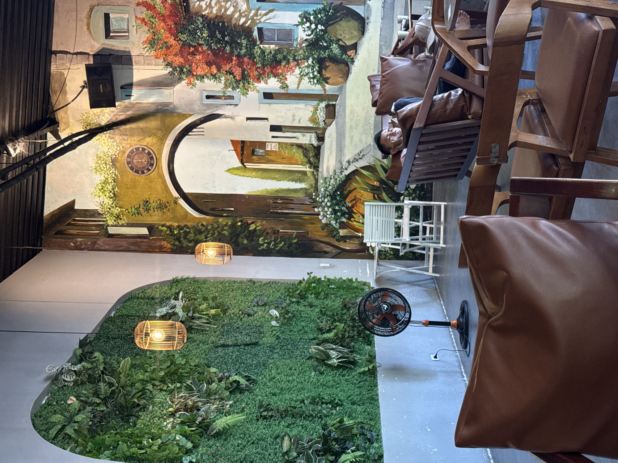 This is an indoor scene at a cafe in Nam Nha Trang Ward, Vietnam. A person is relaxing on a brown leather couch in front of a wall mural depicting a Mediterranean street scene. The mural features an archway, buildings with flowers, and a clock. To the left, a large vertical garden wall is adorned with various plants and two hanging light fixtures resembling birdcages. A black oscillating fan stands on the floor near the garden wall. Several wooden chairs with brown leather cushions are arranged around tables in the foreground.