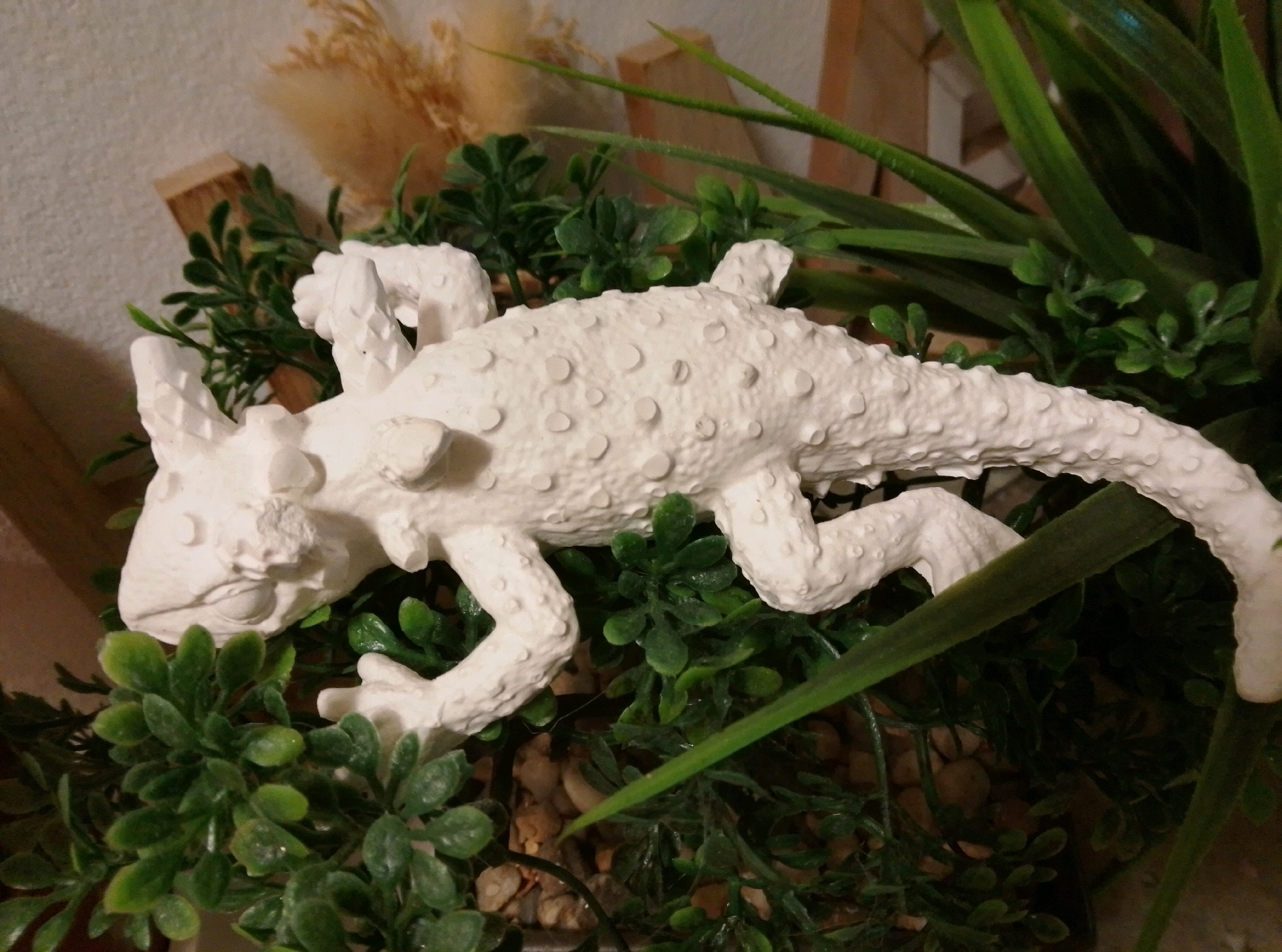The image shows a white, textured sculpture of a lizard positioned among artificial green plants and small stones. The sculpture has a detailed surface with raised bumps and appears to be a decorative item. In the background, there are blurred elements including what looks like a wooden structure and dried plant material. The scene is indoors, likely on a table or shelf, with a light-colored wall in the background. There are no people visible in the image. The location in the city of Wedel, Germany is not discernible from the visual content of the image.