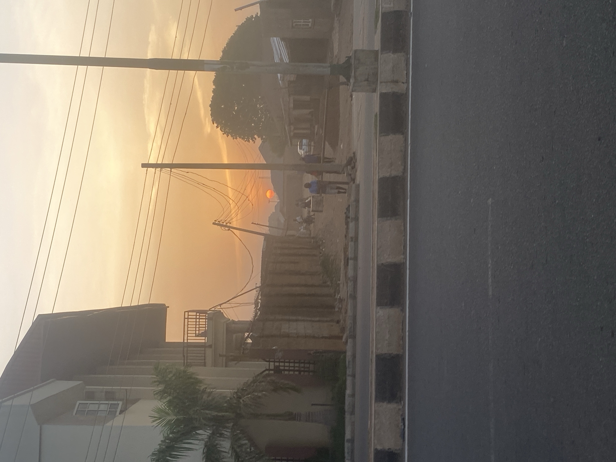 The image depicts an urban scene in Barden Yana, Nigeria, likely at sunset. The sky is a gradient of pale yellow to orange, indicating the time of day. A bright orange sun is visible on the horizon, partially obscured by structures and haze. Overhead power lines are strung across the scene.
Buildings, including a multi-story residence with visible windows and a roof deck, are located on the left side of the frame. Palm trees are present in front of the buildings.
A group of people is situated further down the road, near what appears to be a small commercial structure or roadside stand. Their activities and interactions are not clearly discernable.
A paved road occupies the right side of the image, with a striped curb bordering the walkway. The scene conveys a sense of daily life occurring against a backdrop of a fading sunset.