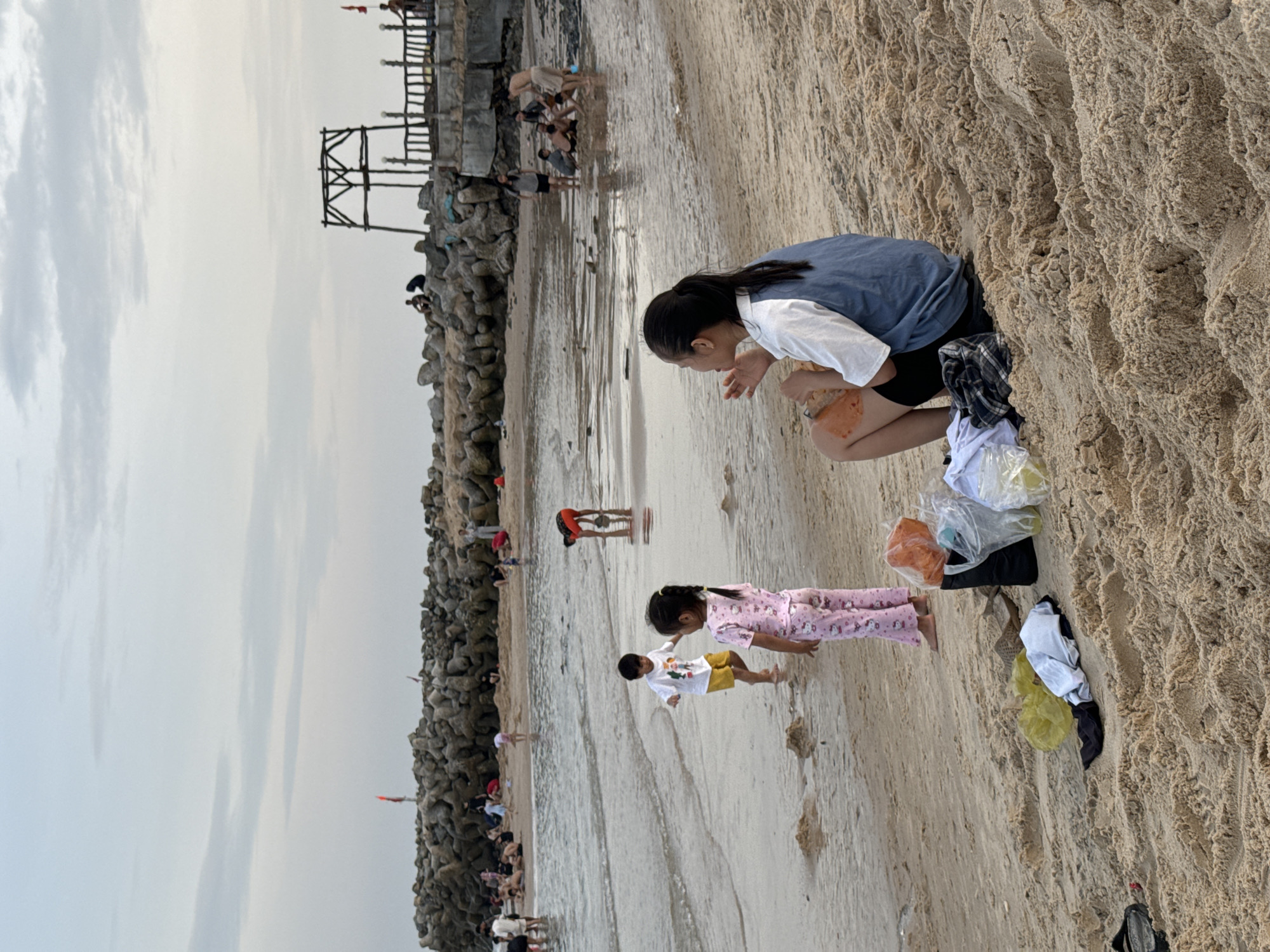 In Phu Thuy Ward, Vietnam, a beach scene unfolds under a hazy sky. In the foreground, a young woman with dark hair tied in a ponytail sits on the sand, wearing a white shirt with a blue vest and black shorts. She is holding a piece of food and appears to be eating. Beside her, on the sand, are various items including a black bag, plastic bags containing what appear to be drinks, a yellow plastic bag, and some folded clothing.
Further towards the water, a young boy in a white t-shirt and yellow shorts is interacting with a young girl in pink pajamas adorned with a cartoon pattern. They are standing on the wet sand near the water's edge. In the background, more people are visible, some sitting on a rocky breakwater, others wading in the shallow water. A wooden structure, possibly a lookout tower or shelter, stands on the breakwater. The overall environment is a beach with sand, shallow water, and a breakwater constructed from large stones. The sky is overcast with light clouds.