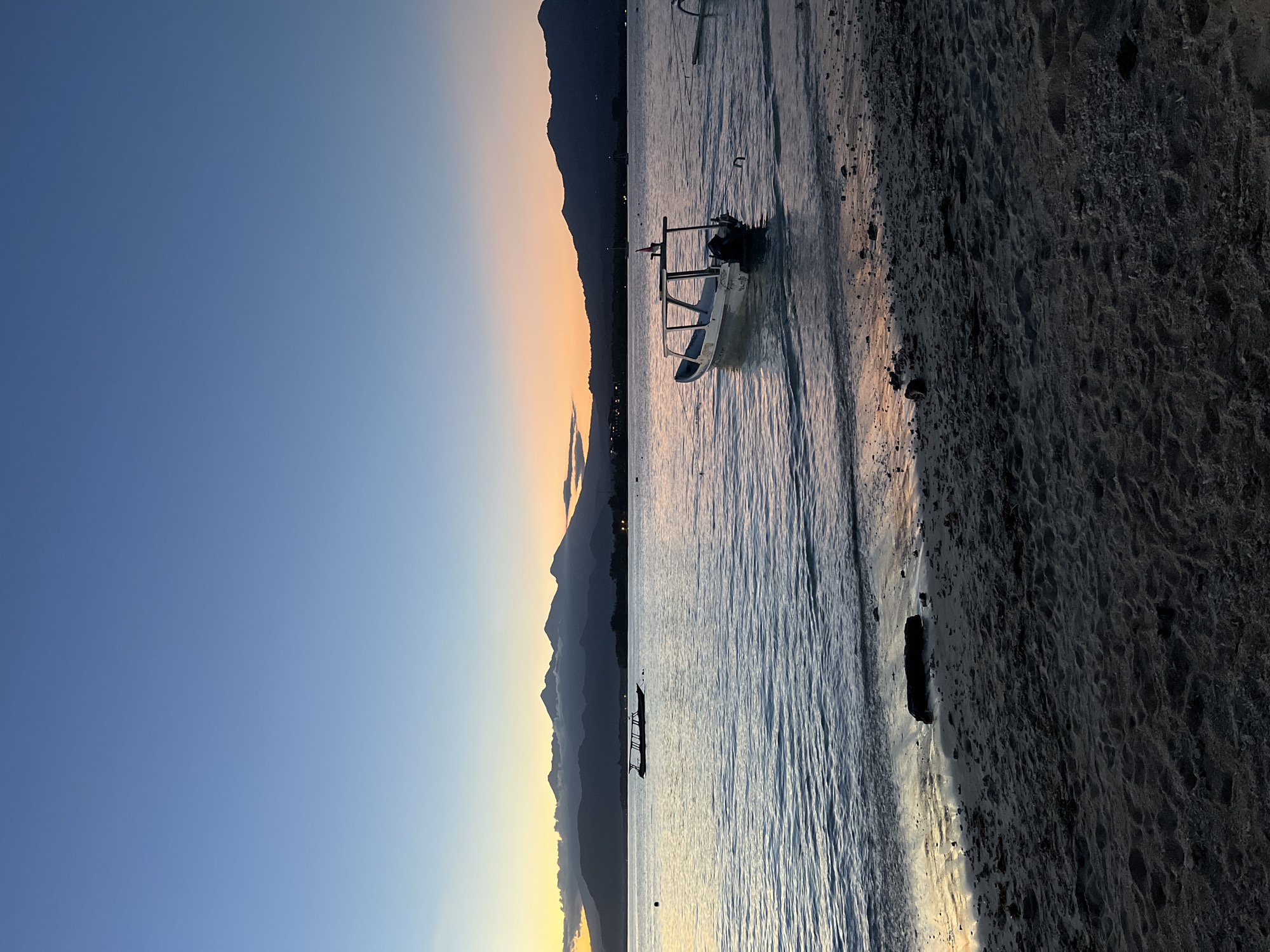 The image depicts a coastal scene at an unknown location in Indonesia, likely during twilight or early morning. The scene is divided vertically; the left side is dominated by a clear blue sky transitioning into a band of orange and yellow near the horizon. Silhouetted mountains are visible against this colorful backdrop.
The right side of the image captures a shoreline. The sea appears calm, with gentle ripples on the surface. Several boats are present; one small boat is close to the shore, and another is further out on the water. A dark, sandy beach stretches along the edge of the water, with varying textures and scattered rocks or debris. The lighting suggests a low-light environment, enhancing the contrast between the sky, mountains, sea, and shore.