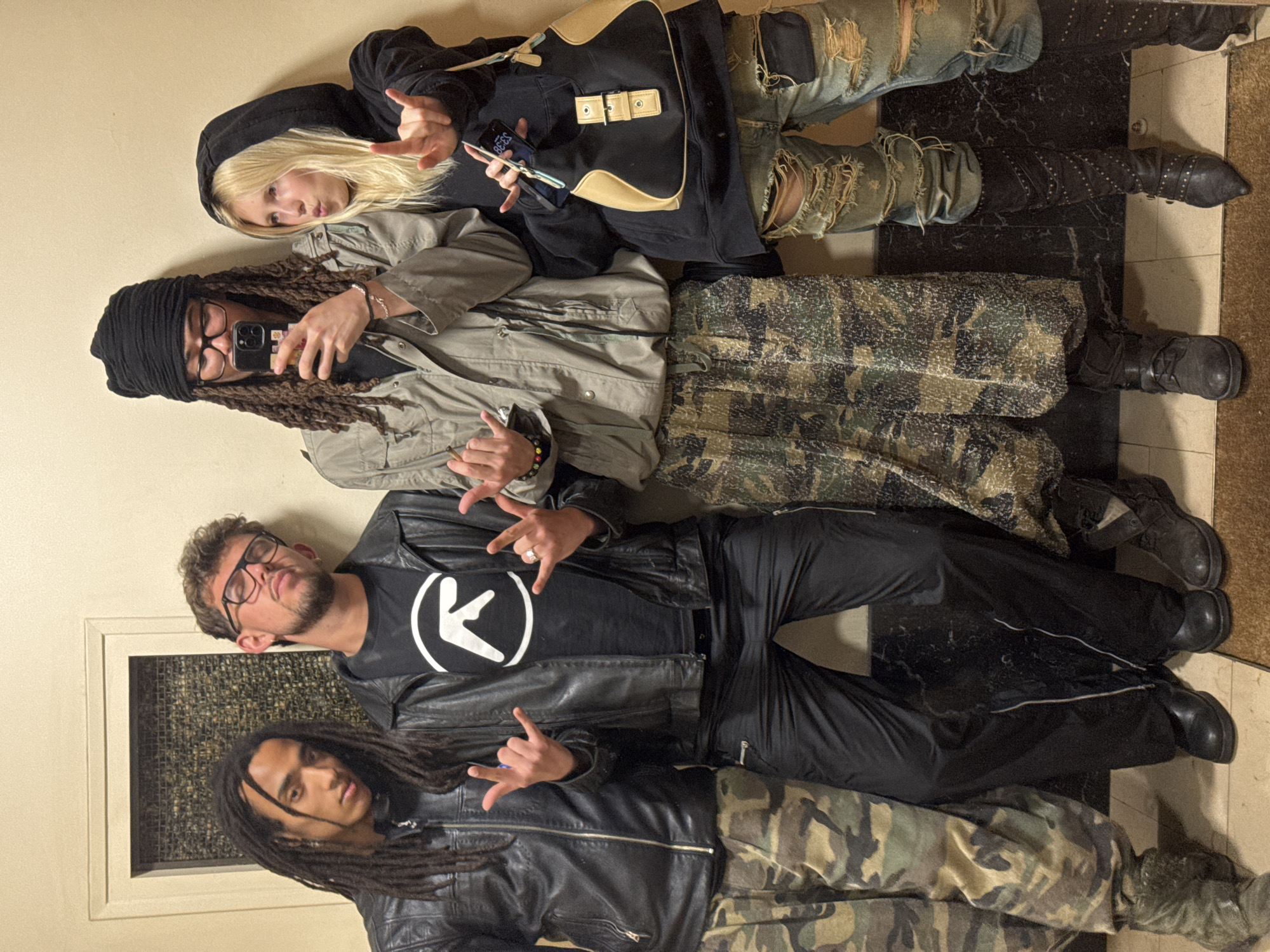 Four individuals are depicted indoors, posing and looking towards the camera. All are making a hand gesture resembling "rock on" or the sign for "I love you" in American Sign Language. From top to bottom:
1. A person with blonde hair wearing a black hoodie and ripped jeans.
2. An individual with dreadlocks wrapped in a black head covering wearing glasses, a light-colored jacket, and camo pants, holding a phone.
3. A man with glasses and a black t-shirt under a black leather jacket and black pants.
4. A person with dreadlocks and a black leather jacket with camo pants.
The background includes a cream wall, a black and white marbled floor, and a decorative element resembling a small window to the left of the frame. There is no indication that this image was taken in Paris, France.