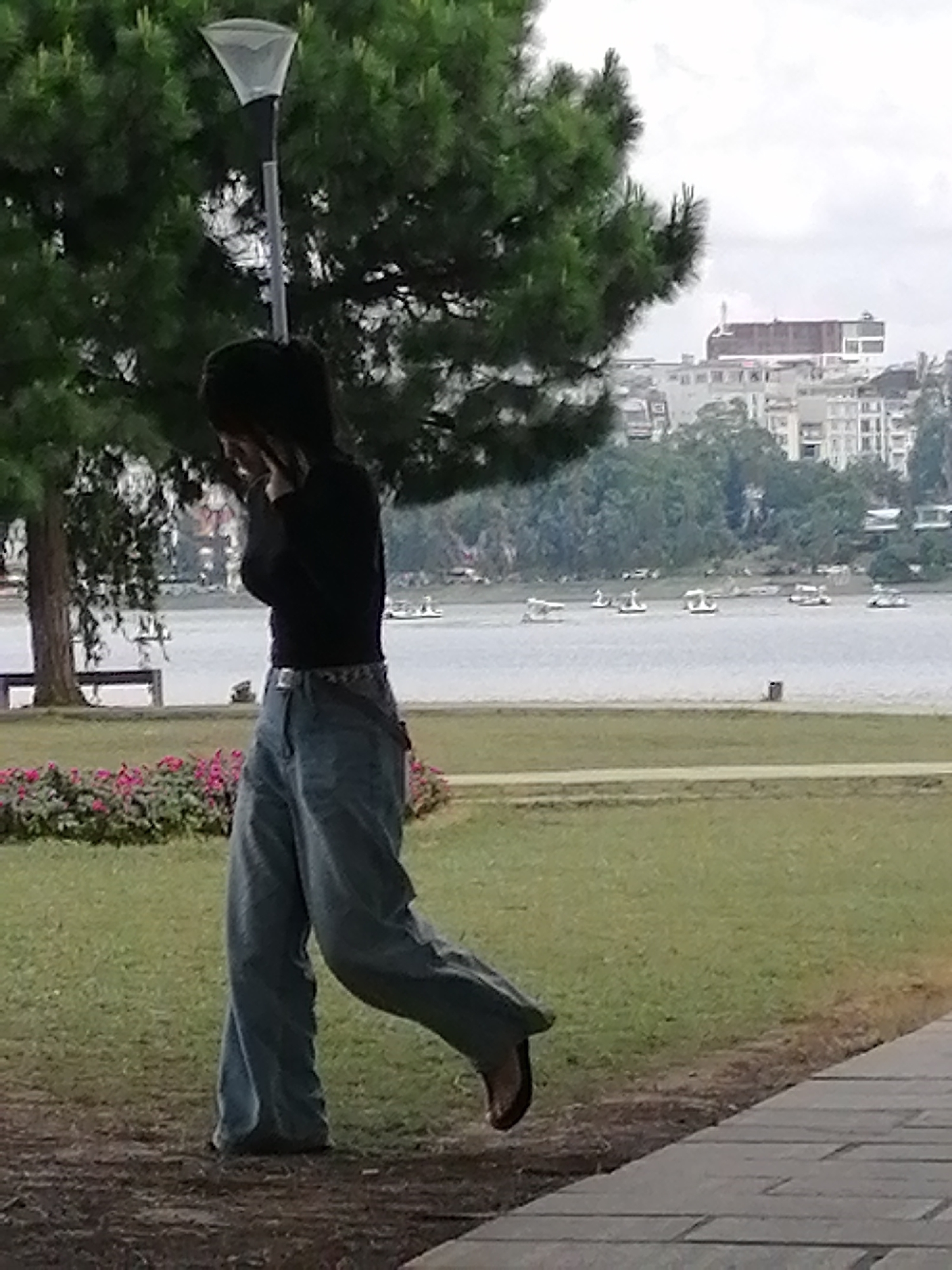 A young person with dark hair, wearing a black long-sleeved shirt and loose-fitting light-wash denim jeans, walks in profile to the right in a park. The person's head is bowed, and their hands are near their face, obscuring it. They are wearing dark flip-flops. The foreground consists of a paved walkway and a grassy area with a line of pink flowers and trees. In the background, a lake or body of water is visible, dotted with several small swan-shaped paddle boats. Across the water, the city of Da Lat, Vietnam, is seen with buildings and trees. A lamppost with a triangular lamp shade is positioned behind the person. The sky is overcast.
