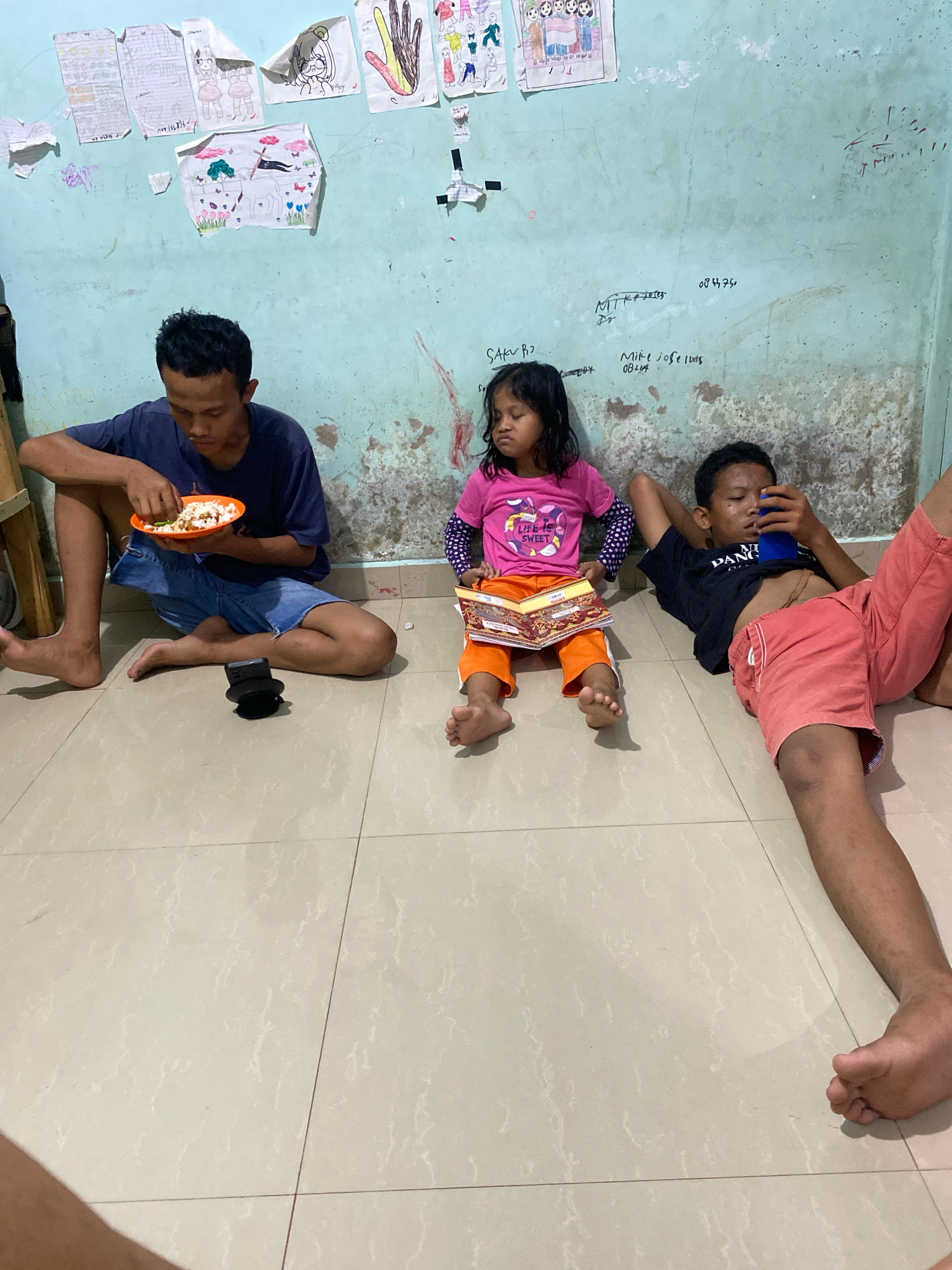 In Batam, Indonesia, three individuals are positioned on a tiled floor in an indoor setting. On the left, a young male sits with his legs extended, eating from an orange plate. In the center, a young female child sits with her legs crossed, holding an open book. To the right, a young male reclines, looking at a blue object, likely a phone, in his hands. The background features a light blue wall adorned with various colorful drawings and some handwritten markings. The floor appears to be made of light-colored ceramic tiles.
