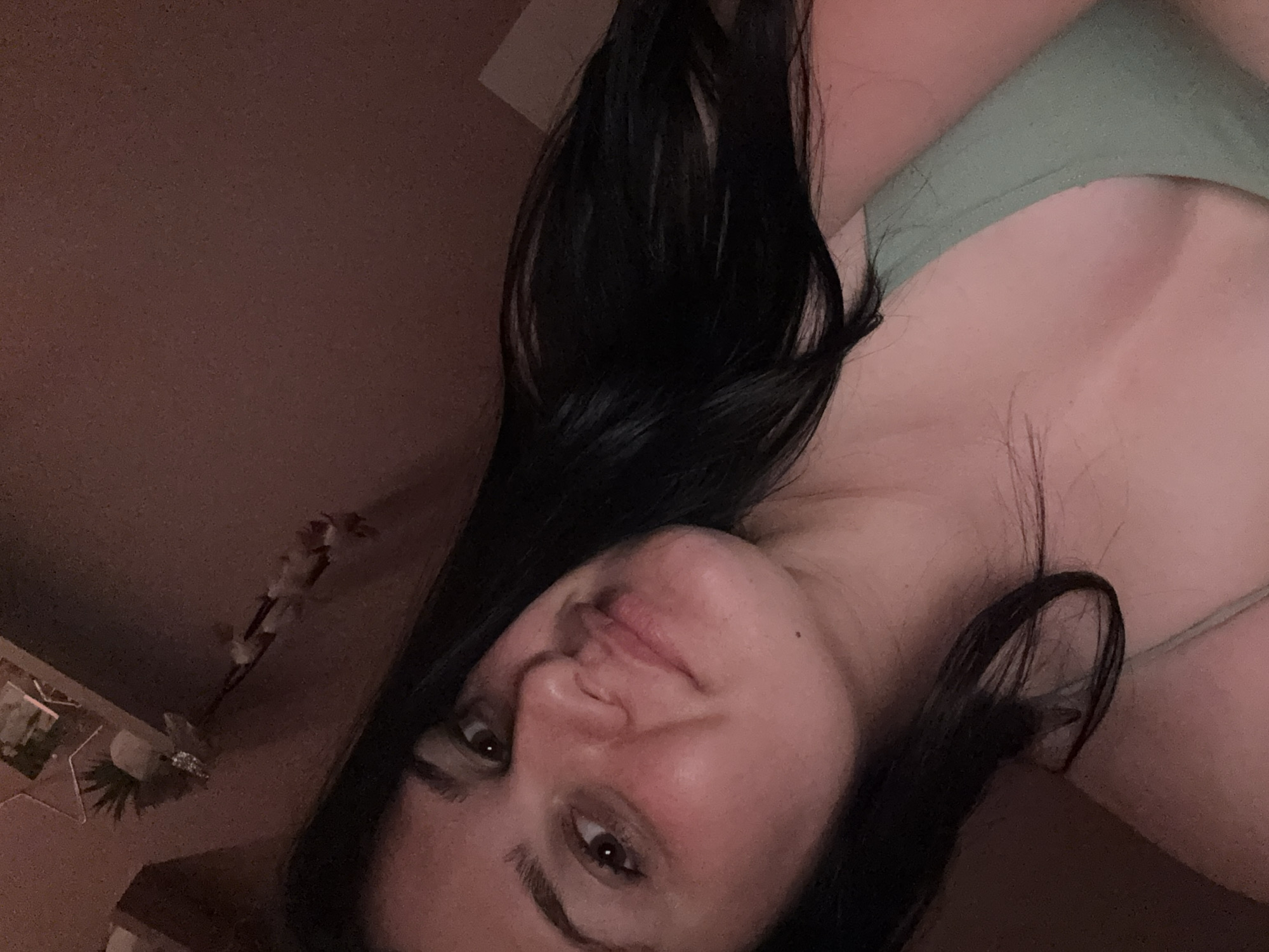A young woman with long, dark hair is captured in a close-up selfie. She is looking towards the camera with a gentle smile. Her skin appears to be flushed, and she is wearing a light green top. The background is dimly lit and appears to be an indoor setting, possibly a bedroom, with a hint of decorative items on a shelf in the upper left corner. There is no indication of the location being in the city of Rogosh, Bulgaria.