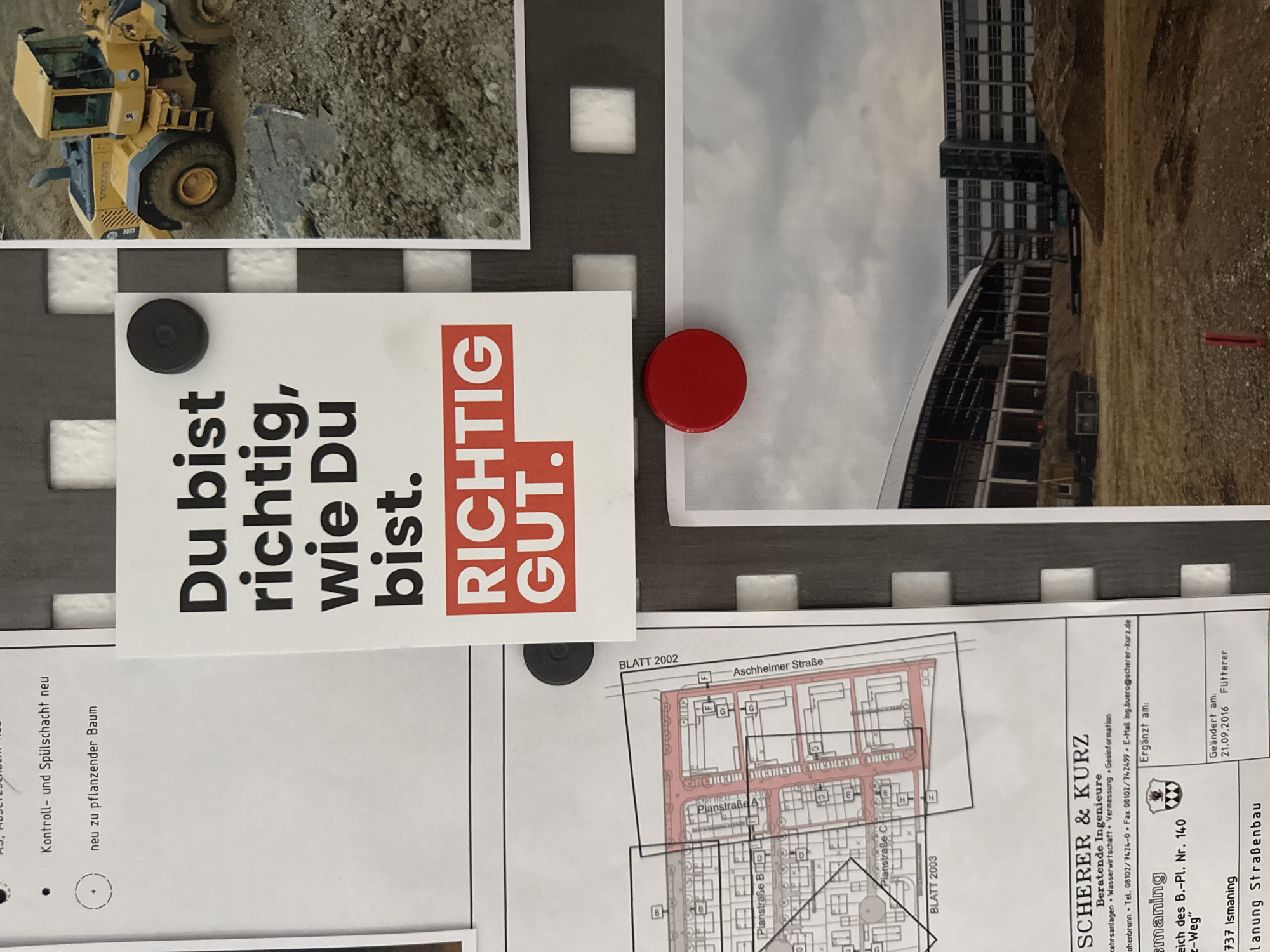 The image depicts a close-up view of a wall or bulletin board with various documents and a large photograph attached. In the upper left, a snippet of a document with German text is visible, mentioning "Kontroll- und Spül-schacht neu" (Control and flushing shaft new) and "neu zu pflanzender Baum" (new tree to be planted).
Dominating the center of the image is a white card with bold German text that reads "Du bist richtig, wie Du bist. Richtig. GUT." (You are right as you are. Right. GOOD.). This card is secured by a black circular magnet.
To the right of the text card, a portion of a photograph shows a yellow Volvo wheel loader on a muddy construction site.
Below the text card and to the left, a blueprint or architectural drawing is visible, showing a grid-like layout with street names like "Planstraße B" and "Planstraße C". The drawing is labeled "BLATT 2002". A red circular magnet is attached to the bottom edge of this blueprint.
Further down, the lower left section of the image displays the letterhead of "SCHERER & KURZ Beratende Ingenieure" (Consulting Engineers), indicating services such as "Verkehrsanlagen • Wasserwirtschaft • Vermessung • Geoinformation" (Traffic facilities, Water management, Surveying, Geoinformation). Contact details include a phone number and email address, with a location potentially linked to Hohenbrunn, Germany, given the text "Hohenbrunn". The document also mentions "Ismaning" and "Ergänzt am:" (Supplemented on:) with a date "21.09.2016".
To the right of the blueprint and letterhead, a large photograph shows a construction site with a partially built structure featuring a pitched roof and multiple stories of windows. Piles of dirt and debris are scattered across the foreground. The sky above is overcast.