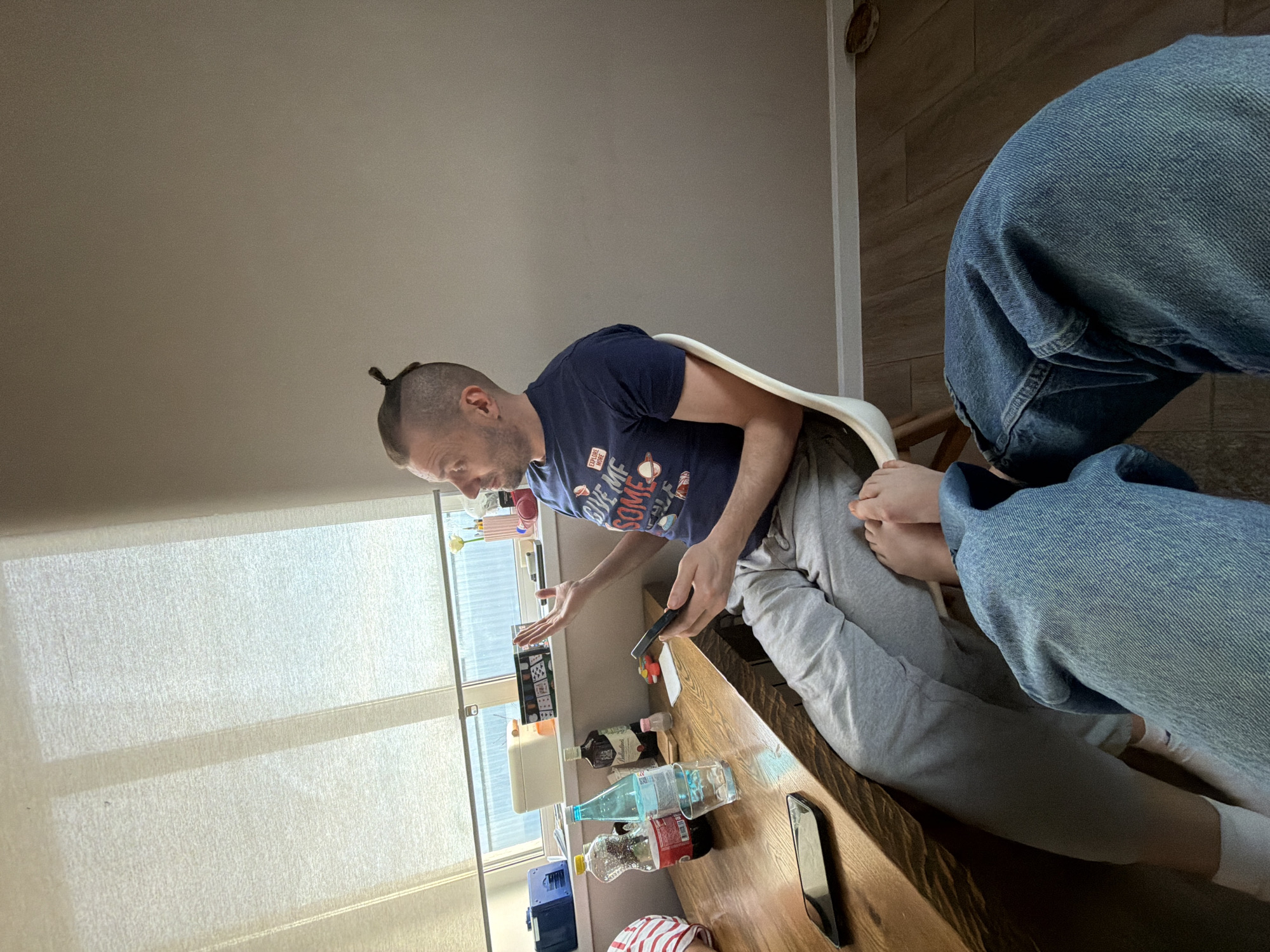 The image shows a man with a partially shaved head and a small ponytail, wearing a dark blue t-shirt and grey sweatpants, sitting in a white chair. He is holding a black object, possibly a phone or remote, and gesturing towards a cluttered wooden table. The table contains various objects including bottles, a white box, and another phone. Another person is partially visible in the lower right corner, wearing blue jeans and white socks. The background includes a window with blinds. The location is unspecified but said to be in the city of Prikamskii, Russia, which is not visible in the image.