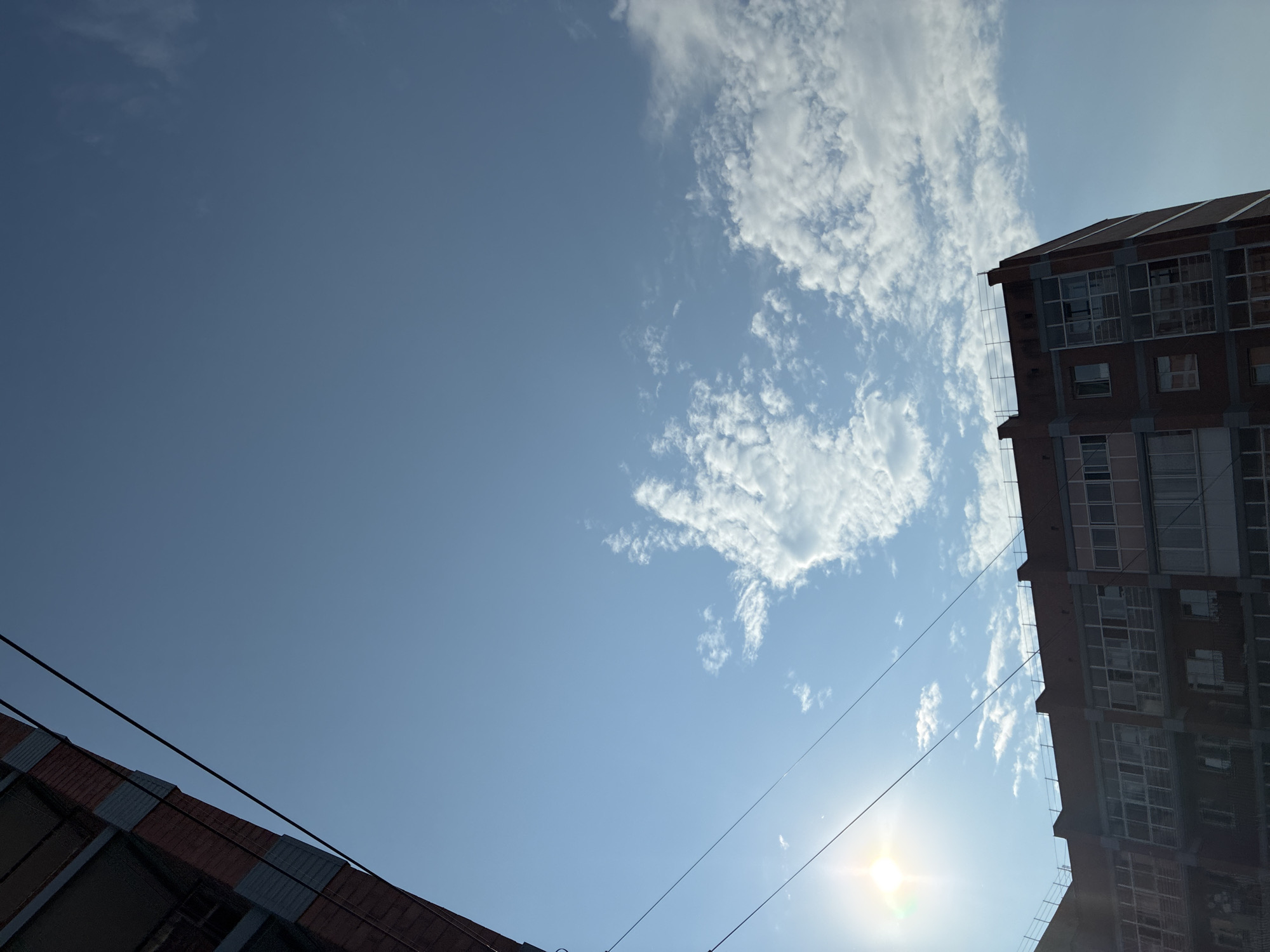 The image shows the facade of a brick apartment building against a bright blue sky with scattered white clouds. The sun is visible in the upper left corner, casting a glare. Two thin, dark wires are stretched across the sky. The top of another building, also brick, is visible in the far upper left corner. The apartment building in the foreground has multiple floors with numerous windows, some with balconies. The overall scene appears to be an exterior urban environment.