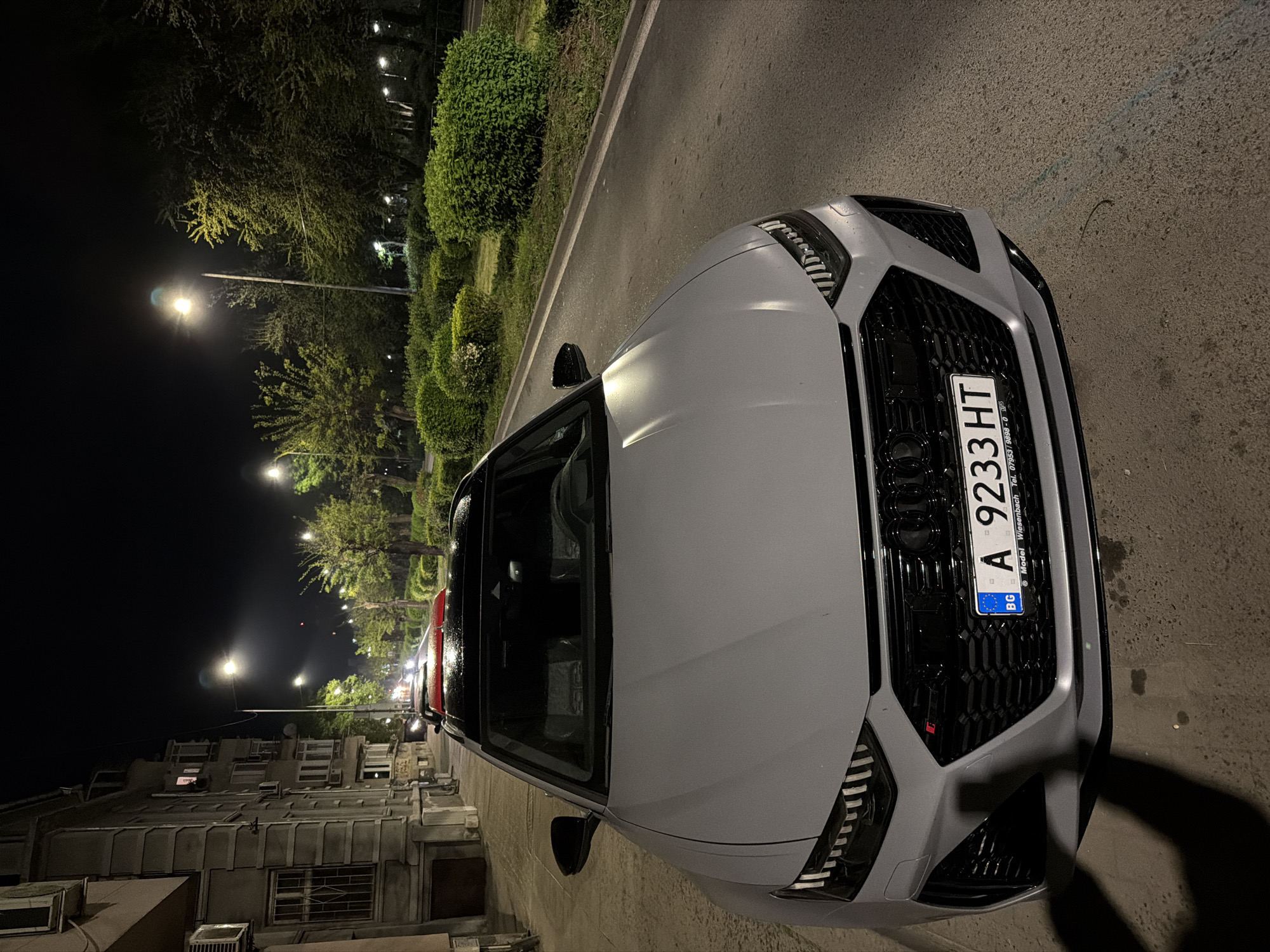 A close-up, night-time photograph shows a light-colored Audi RS e-tron GT parked on the side of a street. The front of the car dominates the view, with the license plate "A 9233 HT" prominently displayed, showing the 'BG' country code for Bulgaria. The street is lined with trees and shrubs on one side, illuminated by streetlights. Buildings are visible in the background. The location is likely Burgas, Bulgaria based on the license plate's country code. The road surface appears to be asphalt. No people are visible.