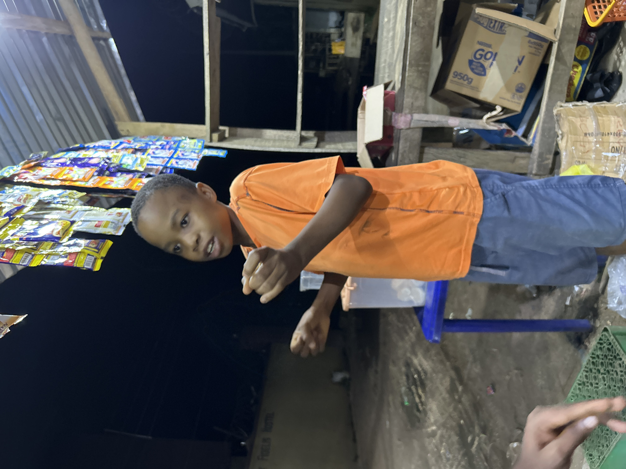 A young boy in an orange shirt and blue shorts stands in front of a small shop in Ihiagwa, Nigeria. The shop has a corrugated metal roof and a wooden structure with shelves displaying packaged snacks and goods. A cardboard box labeled "GO!" is visible on one of the shelves. The ground is dirt and debris. The image is taken at night.