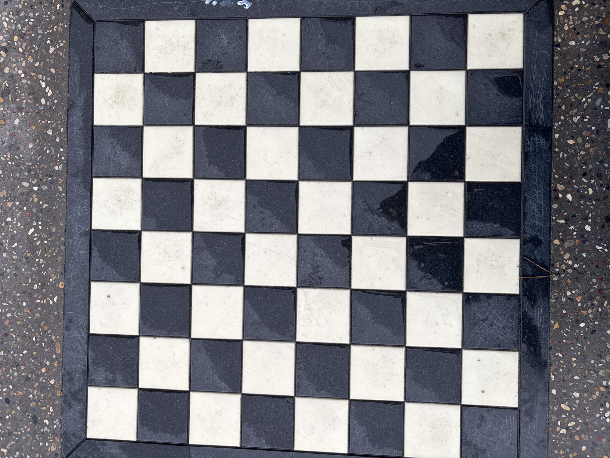 The image shows a weathered, 8x8 black and white tiled chessboard. The board is set on a concrete surface with a speckled pattern, likely an outdoor area. The chessboard has a black frame. The tiles appear to be aged, with scratches and water stains visible on many of the squares. There is no evidence of chess pieces on the board. The image suggests the chessboard is part of a public amenity in the city of Chaikovsky, Russia.