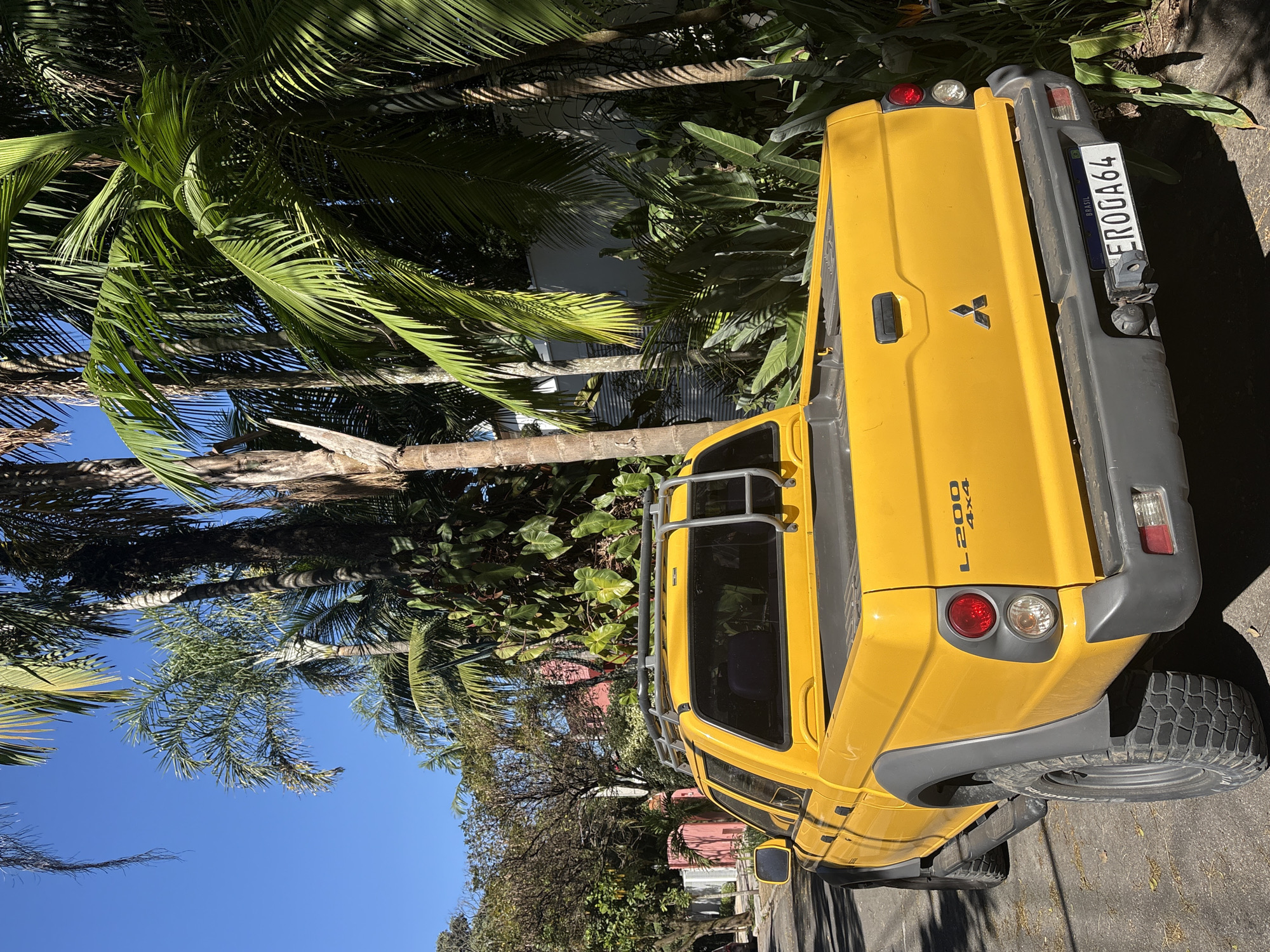 A bright yellow Mitsubishi L200 pickup truck, with "L200 4X4" branding, is parked on a paved street lined with lush tropical foliage, including several palm trees. The truck features a roof rack and a grey utility bumper. Its rear license plate reads "EROGA64" and includes "BRASIL." The scene is illuminated by bright sunlight, casting shadows. The background reveals a red-painted building and more dense greenery. The context suggests a residential area in São Paulo, Brazil, given the license plate and characteristic vegetation. There are no people visible in the image.